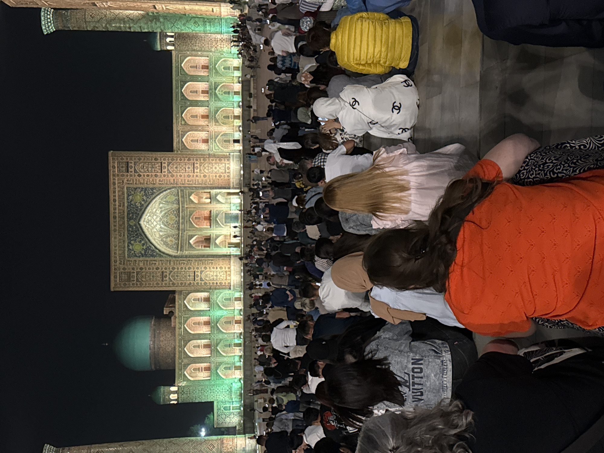 Here is a detailed description of the image:
The image depicts a large group of people praying in front of a grand building. The people are kneeling in rows, suggesting a communal prayer setting. The building in the background is ornately decorated with intricate tilework and illuminated with bright lights, indicating it may be a mosque or a significant religious site.
The location is likely in Samarkand City, Uzbekistan, based on the architectural style of the building, which is characteristic of Islamic architecture found in Central Asia. The presence of the large group of people performing prayer suggests a religious event or a special occasion.