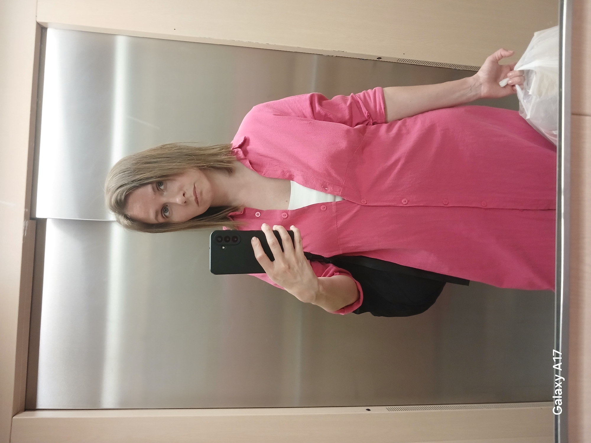 A woman is taking a selfie in an elevator. She is wearing a bright pink collared shirt over a white top, a black bag is slung over her left shoulder, and she is holding a white plastic bag in her right hand. The elevator has stainless steel walls and wood-paneled accents. The bottom of the image shows a reflection of the elevator door and a watermark that reads "Galaxy A17". The woman's expression is neutral. This scene is likely in Yekaterinburg, Russia.