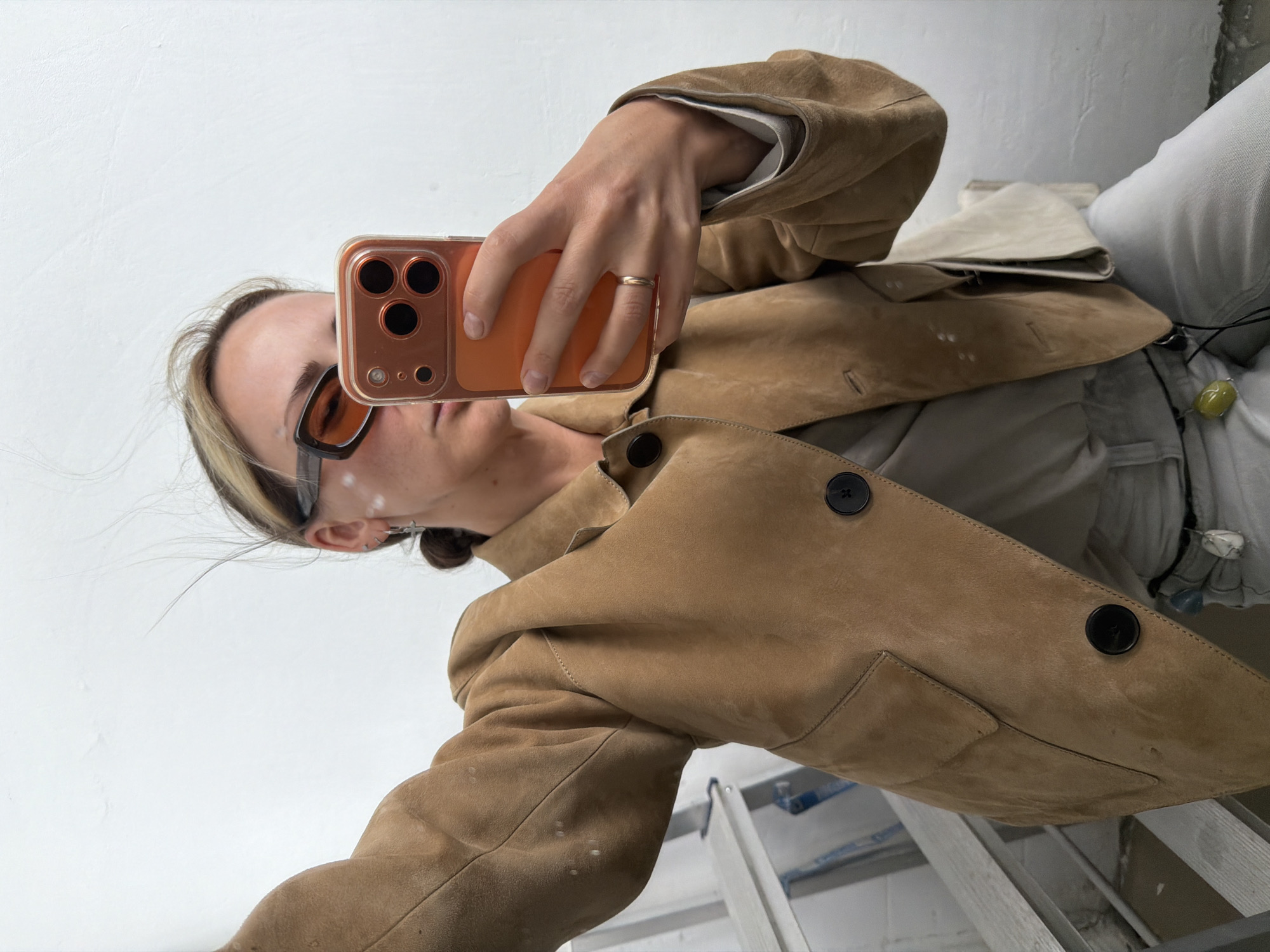 A person is taking a selfie with a smartphone. The phone has a bronze-colored case. The person is wearing sunglasses, a brown jacket with black buttons, and a ring on their finger. There are small dots of what appears to be paint on the side of their face. They are standing on or near a ladder. The background is a white wall. The location is not evident from the image.