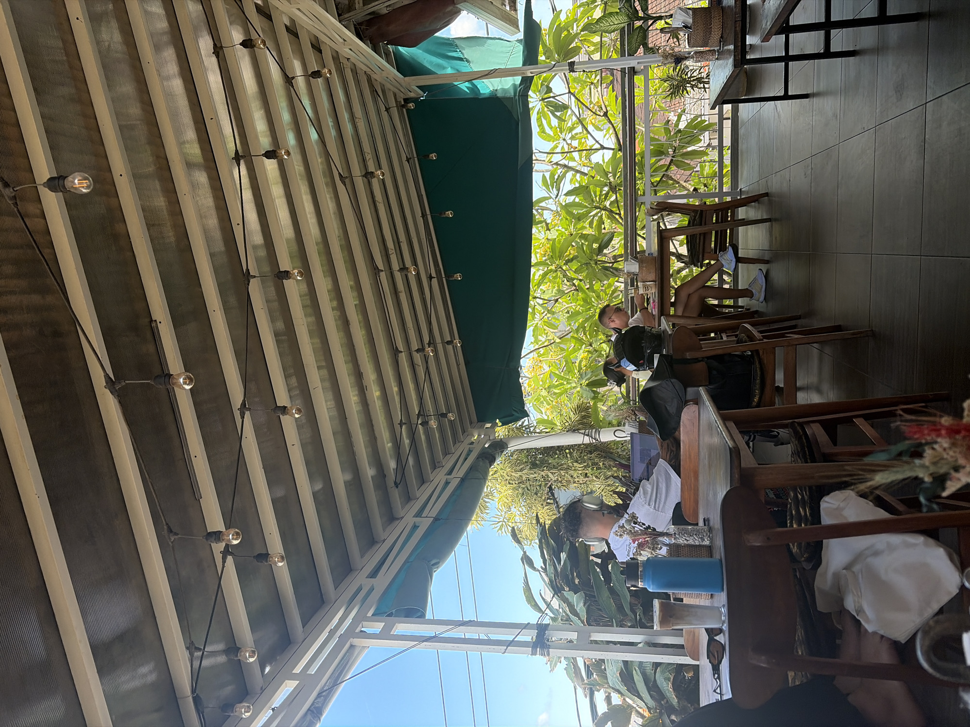 This is an outdoor seating area of a cafe in North Kuta, Indonesia. The scene is captured from a low angle, looking up at the roof structure and out towards lush greenery and the sky. String lights with exposed bulbs hang from the corrugated metal roof, which is supported by white metal beams. A green awning is visible above the railing.
In the foreground, a wooden table holds a glass with an iced coffee, a blue water bottle, and a small vase with dried flowers. Sunglasses rest on the table. Two chairs are positioned at the table. One chair is occupied by a person from behind, wearing a white t-shirt and headphones, looking at a laptop on the table. Another person is seated further back at a table to the right, also looking at a laptop.
The background features dense foliage and trees with vibrant green leaves, suggesting a tropical environment. In the distance, glimpses of rooftops can be seen through the leaves. The floor is tiled with dark grey squares. The overall atmosphere appears relaxed and casual, typical of an outdoor cafe setting in a warm climate.