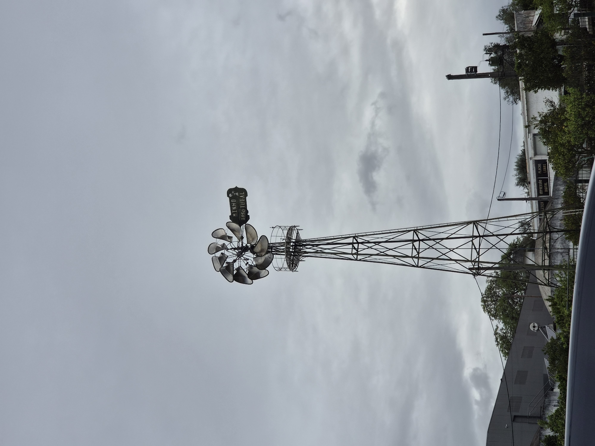 The image shows a decorative windmill in Barcelos, Portugal. The windmill is constructed of metal and features a weather vane at the top displaying "MINHO 1850 TUGAL". The windmill is located near a building with signs visible and a utility pole. The sky is overcast.