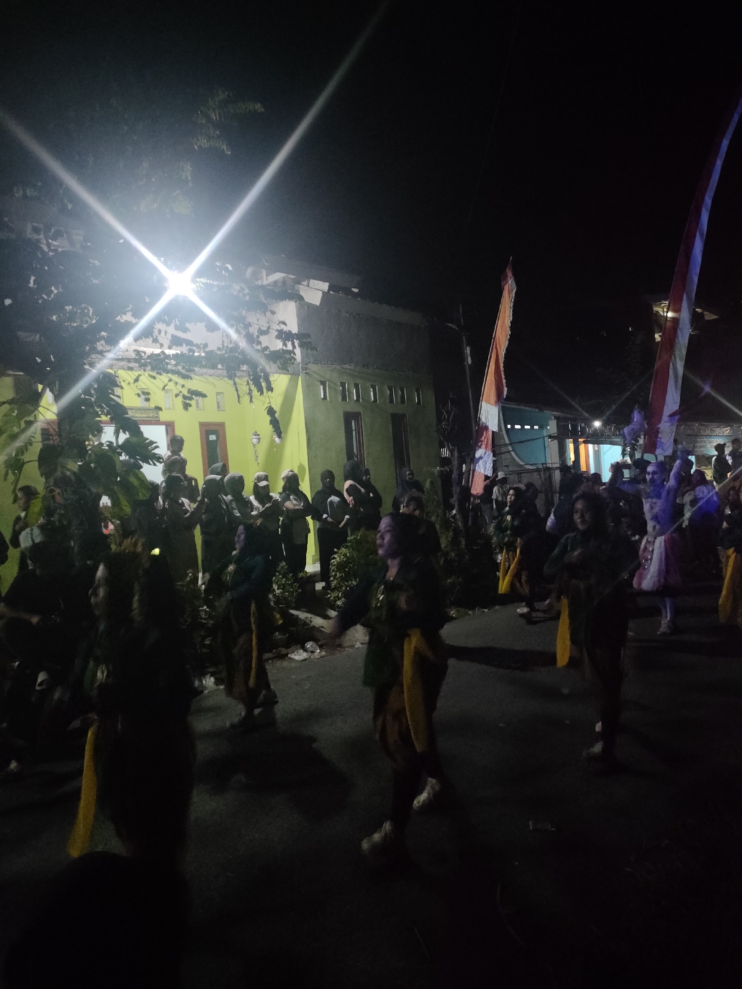 A group of people are gathered on a street in Tongaswetan, Indonesia at night. The scene is illuminated by a bright overhead light, casting strong beams and creating lens flares. Many individuals are wearing traditional clothing, including elaborate headdresses and patterned fabrics. Some are holding or displaying flags. The architecture visible in the background suggests a residential area, with a light-colored building and some greenery. The people appear to be participating in a celebration or performance, with some in motion, suggesting dancing or marching. The ground is paved and appears to be a street. The overall atmosphere is festive and communal.