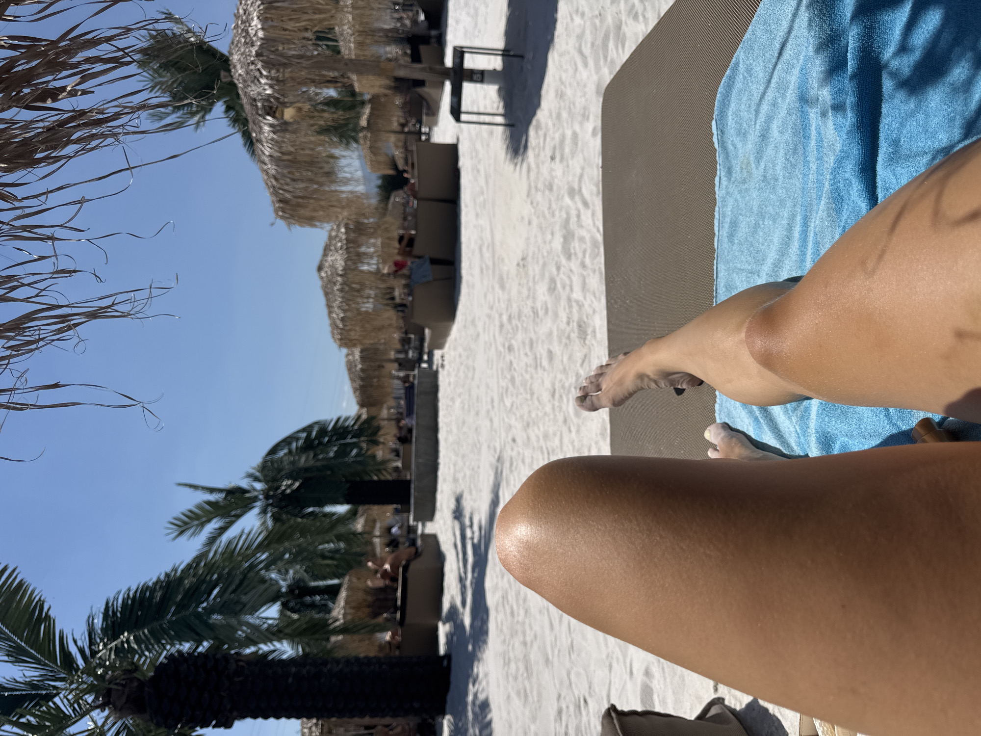 The image depicts a first-person perspective of someone relaxing on a sun lounger at a beach in Kazichene, Bulgaria. The foreground features a person's legs, with a blue towel draped over the lounger. The sand appears white and fine-grained. In the background, several thatched umbrellas and palm trees are visible, suggesting a resort or a beach club setting. A few people are sparsely scattered in the background, seated or lounging under the umbrellas. The sky is clear and blue, indicating a sunny day. The scene conveys a sense of leisure and relaxation in a tropical or Mediterranean-like environment.