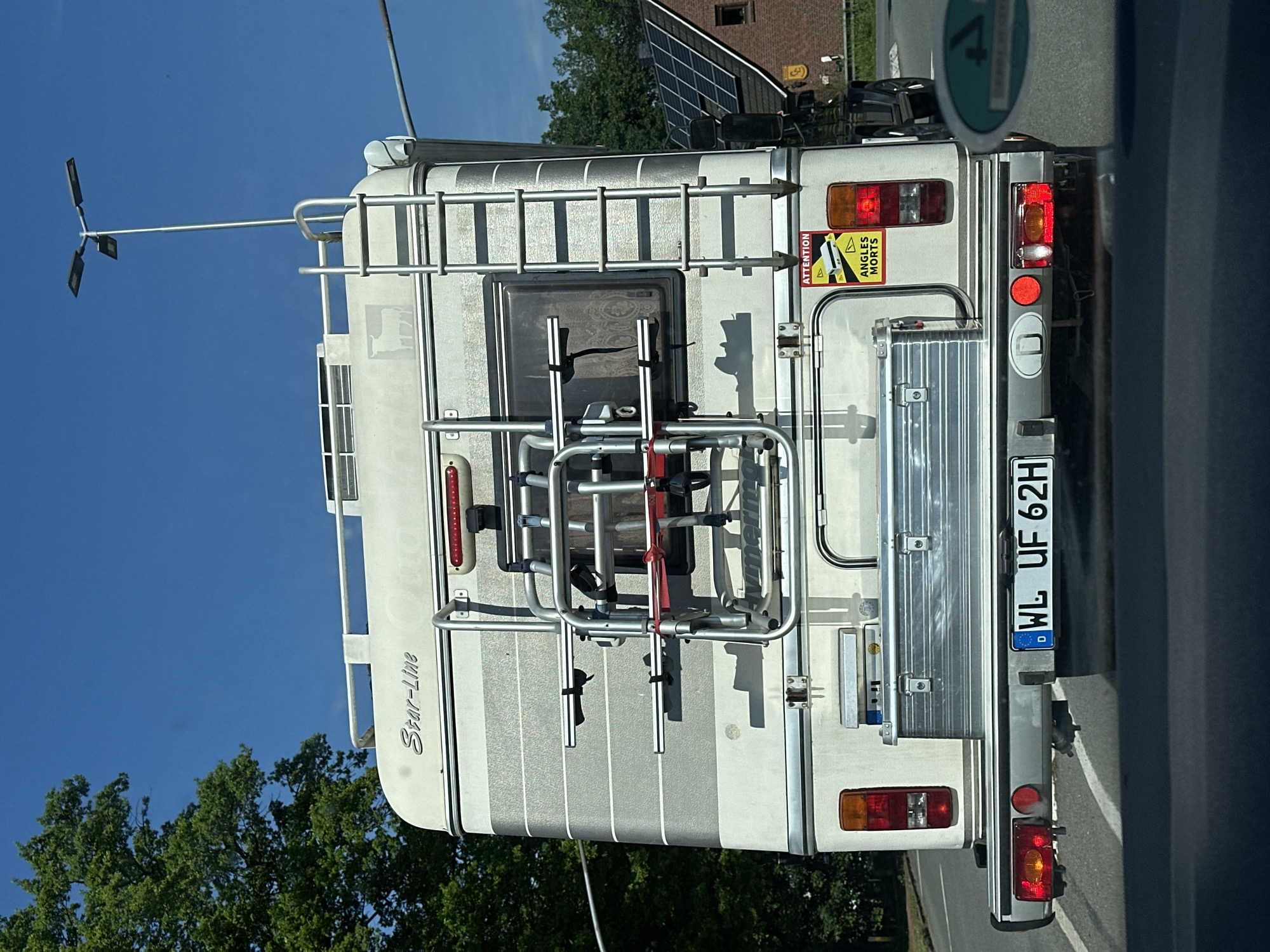 The image shows the rear of a "Star-Line" camper vehicle, with German license plate "WL UF 62H." The vehicle has a ladder leading to the roof, a bicycle rack mounted at the rear, and a warning sticker "ATTENTION ANGLES MORTS". The vehicle is located on a road in Neu Wulmstorf, Germany.