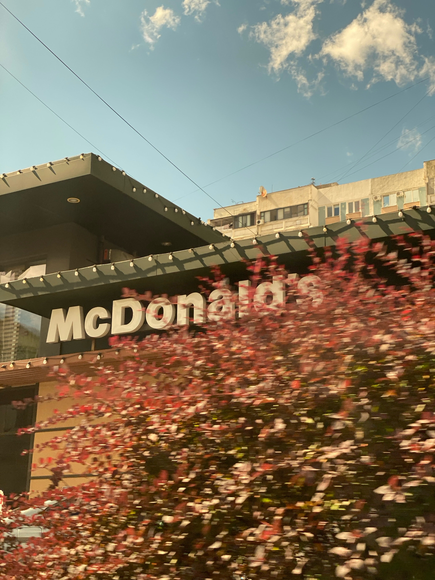 The image shows a McDonald's restaurant in Chișinău, Moldova. The McDonald's sign is prominently displayed on the building. In the foreground, there is a blurry image of red-leaved trees, suggesting motion or a fast shutter speed. The sky is blue with some clouds. In the background, there are apartment buildings. The overall scene appears to be a daytime shot taken from a moving vehicle.