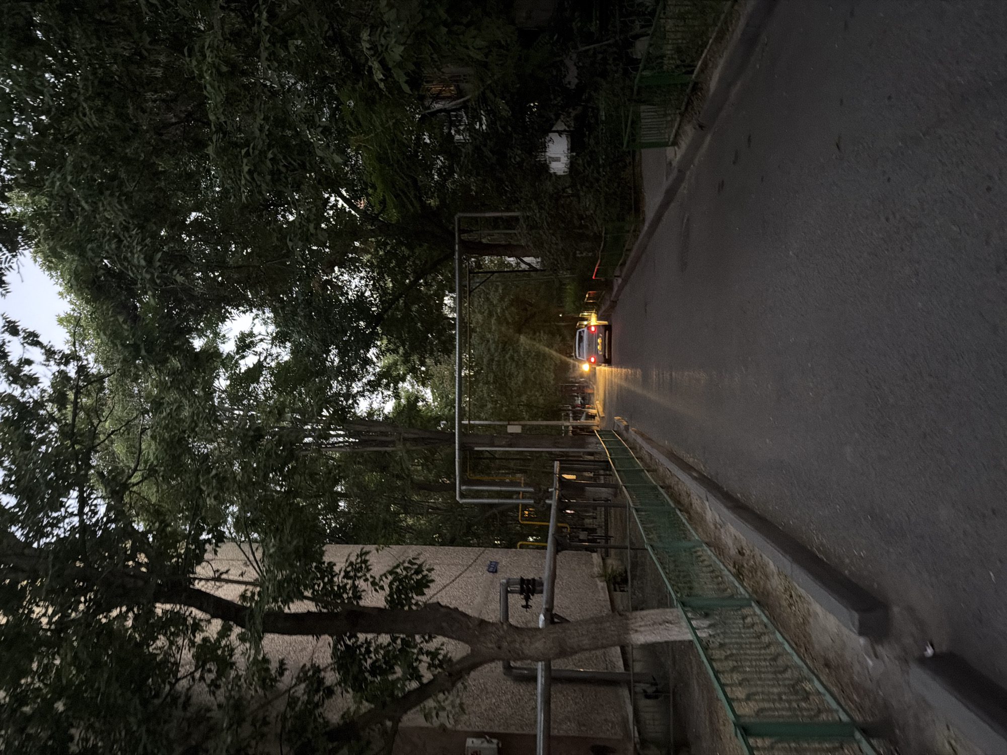 A gray sedan with its brake lights illuminated drives down a paved road lined with trees and a green metal fence on the left. The scene appears to be in a residential area of Shymkent, Kazakhstan, possibly an inner courtyard of an apartment complex, given the presence of external pipes on the side of a building on the left and the overall architecture. The sky is overcast and the lighting suggests it is either early morning or late evening.
