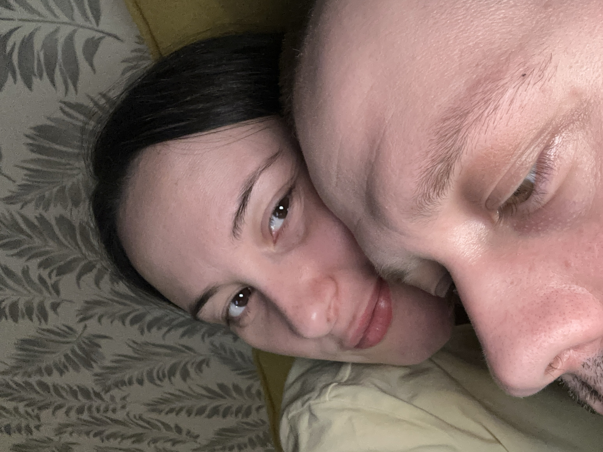 A close-up photograph depicts two individuals, a woman and a man, lying closely together. The woman has dark hair and is looking at the camera with brown eyes. The man is bald and appears to be leaning his head against the woman. The background includes a patterned surface that seems to be a wall or a piece of furniture and a yellow blanket or sheet. The location and city (Chaikovsky, Russia) cannot be verified from the given image.