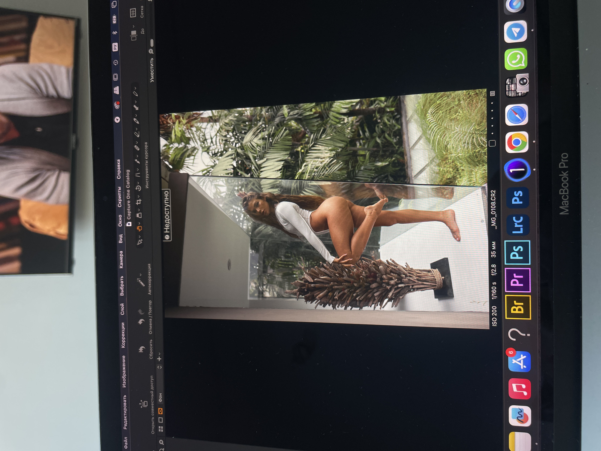 The image is a photo of a MacBook Pro screen displaying an open Capture One application. Within the application, a photograph of a woman in a white bodysuit is being edited. She is posing outdoors, leaning on a decorative arrangement of sticks/twigs on a stand, with lush greenery visible in the background. The laptop screen also shows the Capture One editing interface, the dock with various application icons, and the MacOS menu bar. The location (Kazan, Russia) is not apparent from the image.