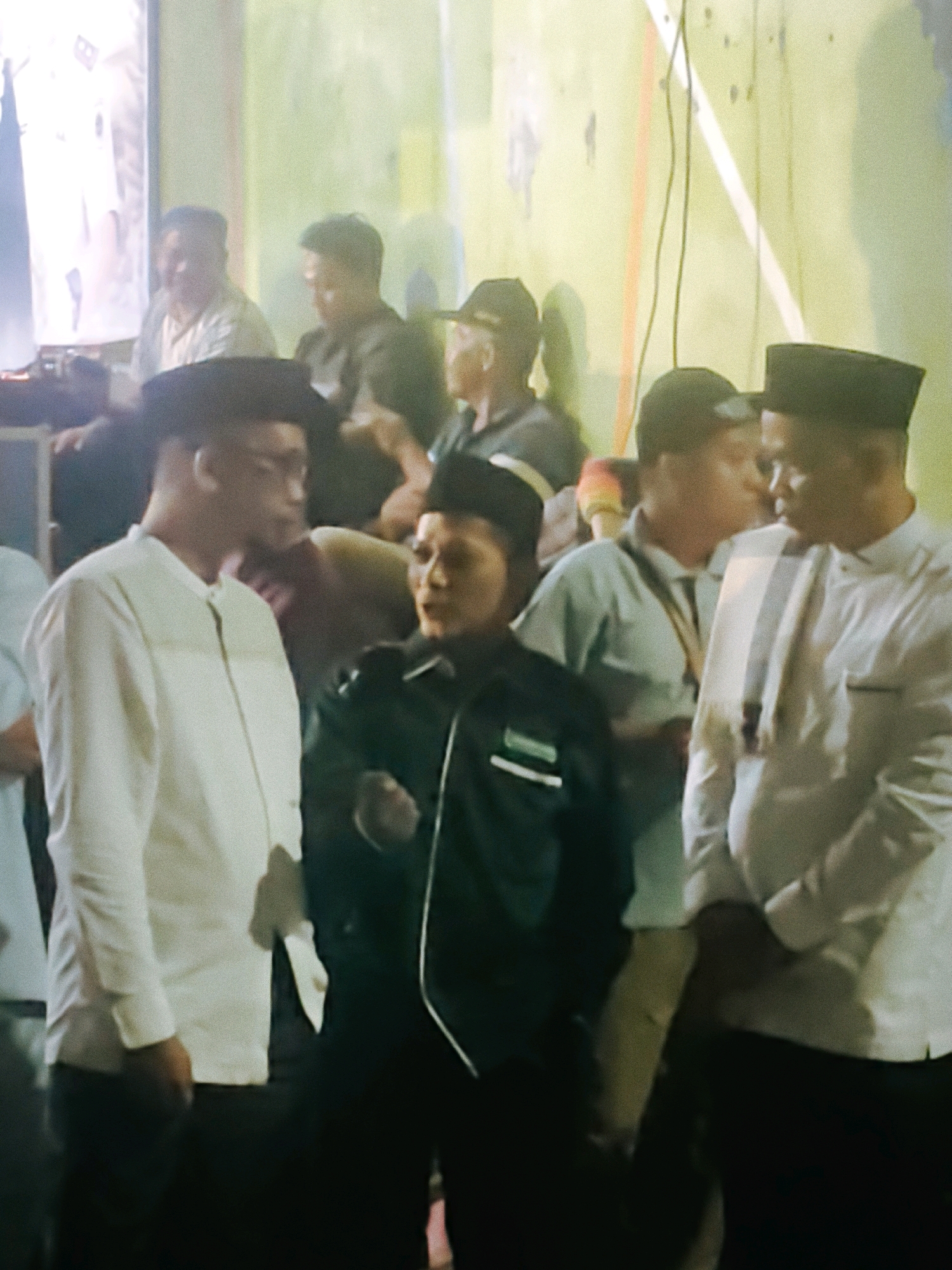 In Rengat, Indonesia, a group of men are gathered outdoors, possibly during an event. The foreground features a man in a white long-sleeved shirt and dark pants, facing to the right. To his right, another man in a dark jacket and a black cap is also facing right, with his hands clasped. Further back, several other men are visible, some wearing caps. The background is a light-colored wall with vertical and diagonal streaks of orange and white. The lighting appears to be artificial and is uneven, casting shadows.