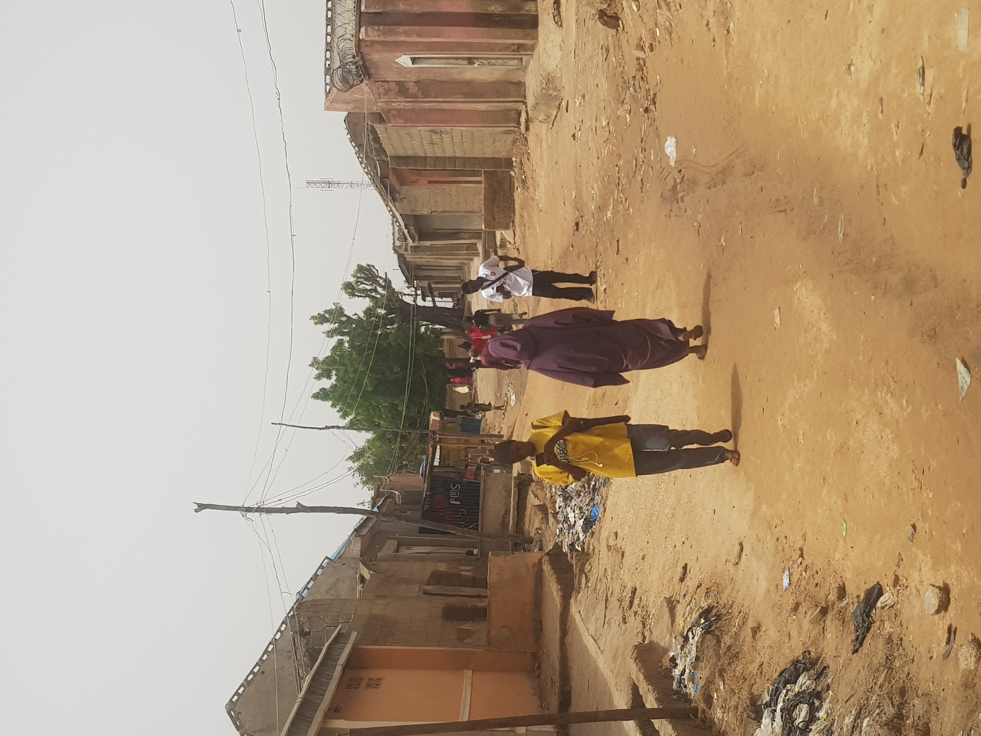 The image depicts a dusty street in Dabawa, Nigeria. The environment appears arid, with buildings constructed from weathered materials. Three individuals are present: one in a yellow shirt, one in a purple head covering and dress, and one in a white shirt. Garbage and debris are scattered across the ground.