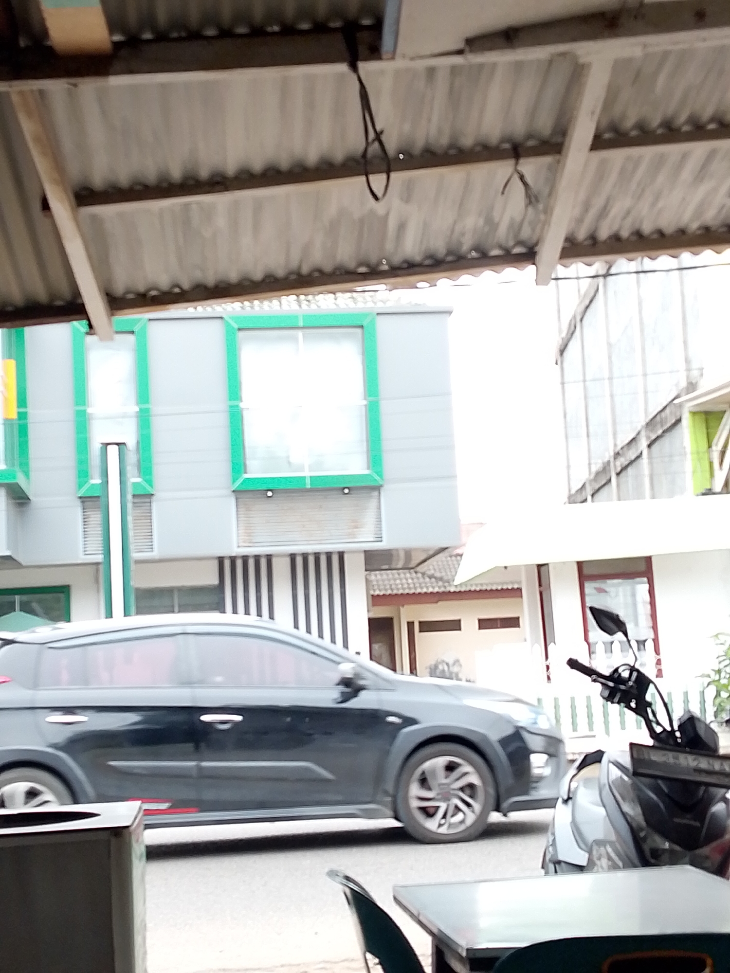 The image depicts a street scene in Lhokseumawe, Indonesia, focusing on a black car in motion, possibly a hatchback, with tinted windows and chrome door handles. The car is traveling on a paved road alongside a row of buildings with modern architecture featuring grey facades and bright green window frames. To the right, a silver and black motorcycle is parked. The foreground shows a glimpse of a table and chairs, suggesting an outdoor seating area, possibly at a cafe or restaurant. The overhead view reveals a corrugated metal roof structure. The overall impression is a snapshot of everyday urban life.