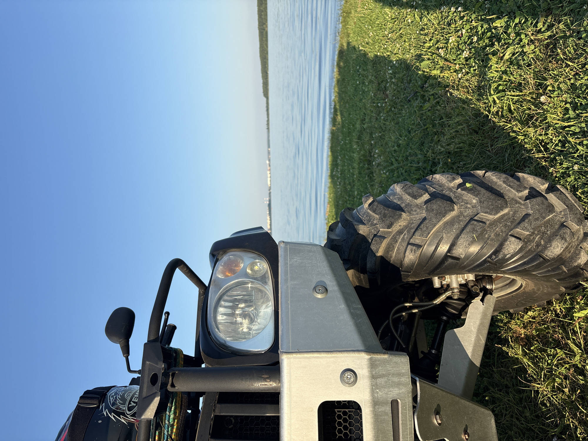 The image shows a close-up, low-angle view of the front of a black and silver ATV parked on a grassy area next to a calm body of water. The ATV has a large, rugged tire on the right side, a silver bumper, and a visible headlight. The handlebars and a helmet are partially visible at the top left. The background consists of a serene blue sky meeting a wide expanse of water, with a distant shoreline featuring trees and industrial structures. The lighting suggests it is either early morning or late afternoon. The location is identified as Konets-Bor, Russia.