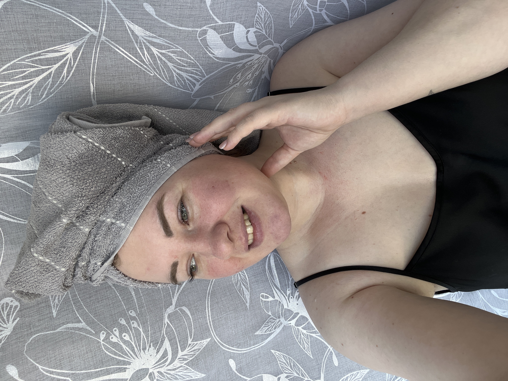 A close-up, selfie-style photograph depicts an adult female in Chaikovsky, Russia. Her head is enveloped by a gray terrycloth towel patterned with white dotted lines forming a grid. She possesses fair skin, exhibiting areas of redness on her cheeks, jawline, and upper chest. Her green eyes are directed towards the left, and a subtle smile is present on her face, partially revealing her upper teeth. She is dressed in a black top with thin spaghetti straps, exposing her right shoulder and parts of her upper chest. Her left hand, featuring lightly polished fingernails, is positioned with fingers gently splayed against the left side of her neck and jawline. A small, dark, triangular tattoo is visible on her right forearm. The background consists of a light gray fabric, likely a curtain or wallpaper, adorned with a repeating pattern of white outlined floral and leaf motifs.