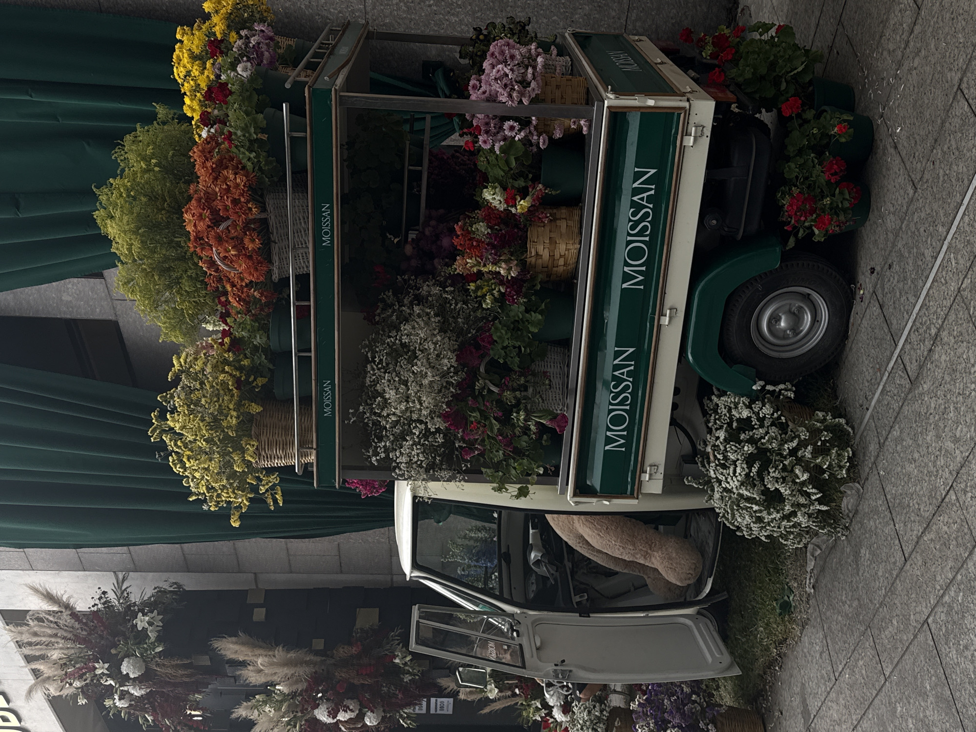 A stationary "Moissan" branded flower truck is parked on a city street. The truck bed is overflowing with assorted flowers and greenery. The open door reveals a large teddy bear inside the cab. Additional floral arrangements surround the base of the truck and adorn the storefront in the background. The location is determined to be Mexico City, Mexico.