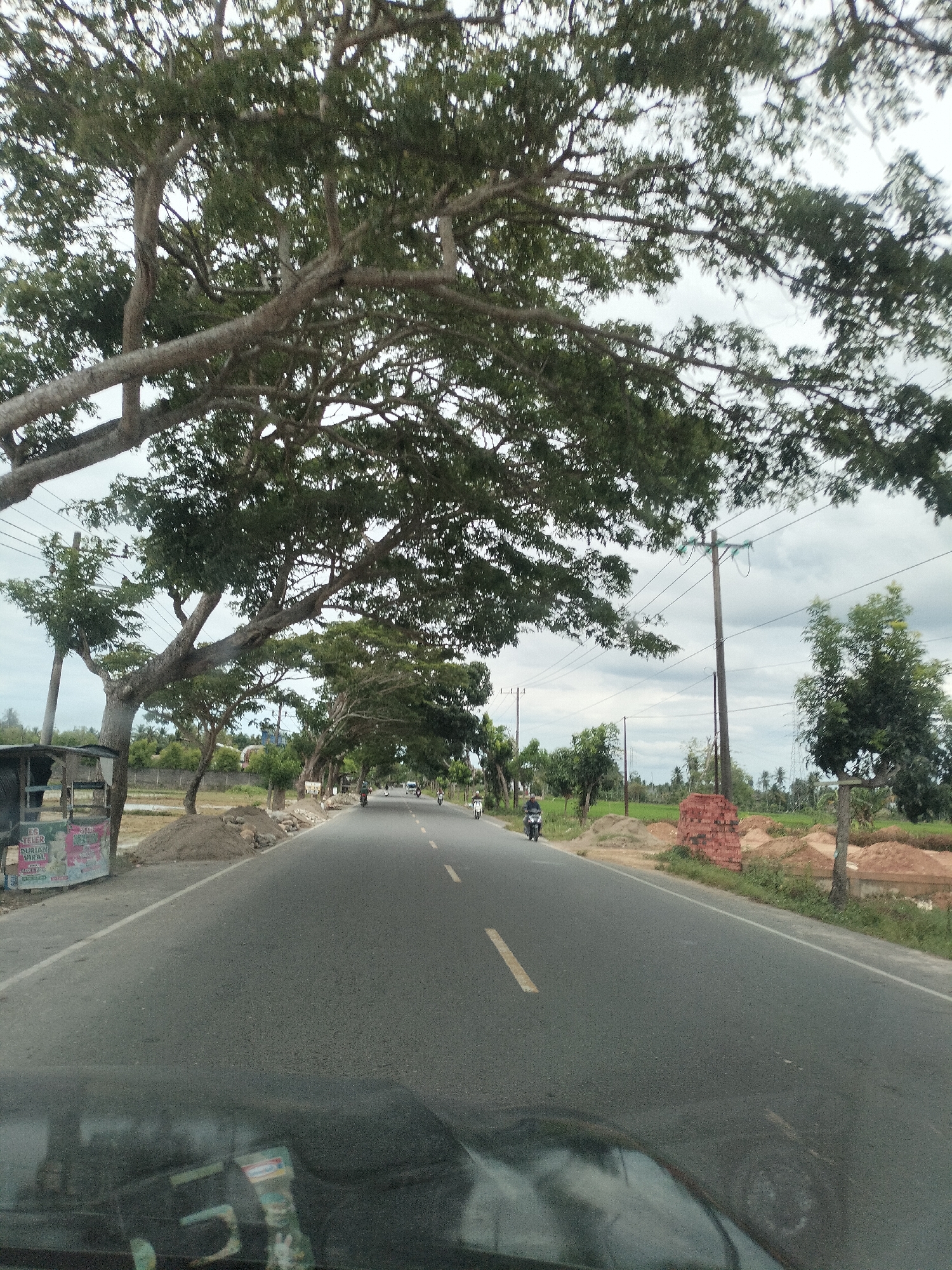 The image depicts a road scene in Putoh Sa, Indonesia, characterized by a paved asphalt road with a white dashed line marking the lane. Large trees with dense foliage arch over the road from the left side. On the left side of the road, there is a small roadside stall or kiosk with signage, and a pile of dirt and construction materials. Several utility poles with electrical wires run along the right side of the road. A few motorcycles are visible on the road, with riders present on two of them. In the distance, more trees and vegetation can be seen, along with cloudy sky. The foreground shows the reflection on what appears to be a car's windshield or dashboard.