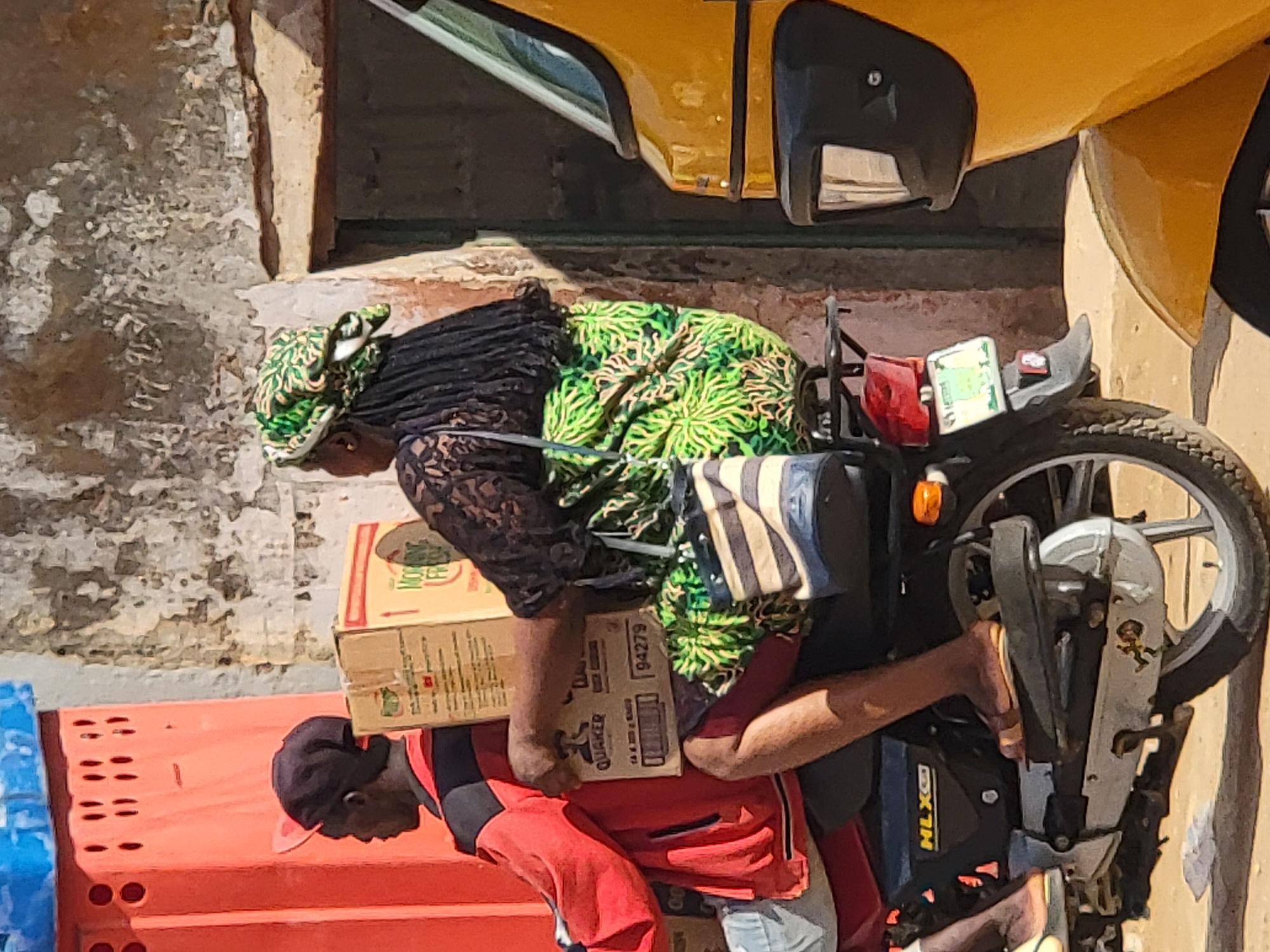 This photograph captures an outdoor scene, likely a street or market in Maiburiji, Nigeria, under bright daylight. The main subjects are a man and a woman engaged in what appears to be a loading or unloading activity involving a motorcycle and several cardboard boxes.
The woman is positioned towards the rear of a black motorcycle, possibly sitting on it or standing very close. She wears a vibrant green and yellow patterned headwrap and dress over a dark, lace-patterned long-sleeved top. A striped black and white bag hangs near her.
The man, with dark skin, is seen bent over, facing away from the camera, wearing a dark baseball cap with a red brim, a red and black shirt, and light-colored pants. He is handling cardboard boxes, suggesting he is loading them onto the motorcycle or moving them from the vehicle. One visible box on the woman's lap/leg has "QUAKER" and the number "94279" printed on it. Another box shows partial text "MORNA" and "TEE".
The motorcycle is black with a red taillight and a partially visible license plate. The brand "HLX" is visible on its side. In the background, part of a yellow vehicle (possibly a car or tricycle) is visible. To the left, bright orange and blue plastic crates are stacked. The overall setting includes a worn, light-colored building facade, suggesting an urban or semi-urban environment. The strong shadows indicate a sunny day. The scene depicts everyday commercial activity or transport.