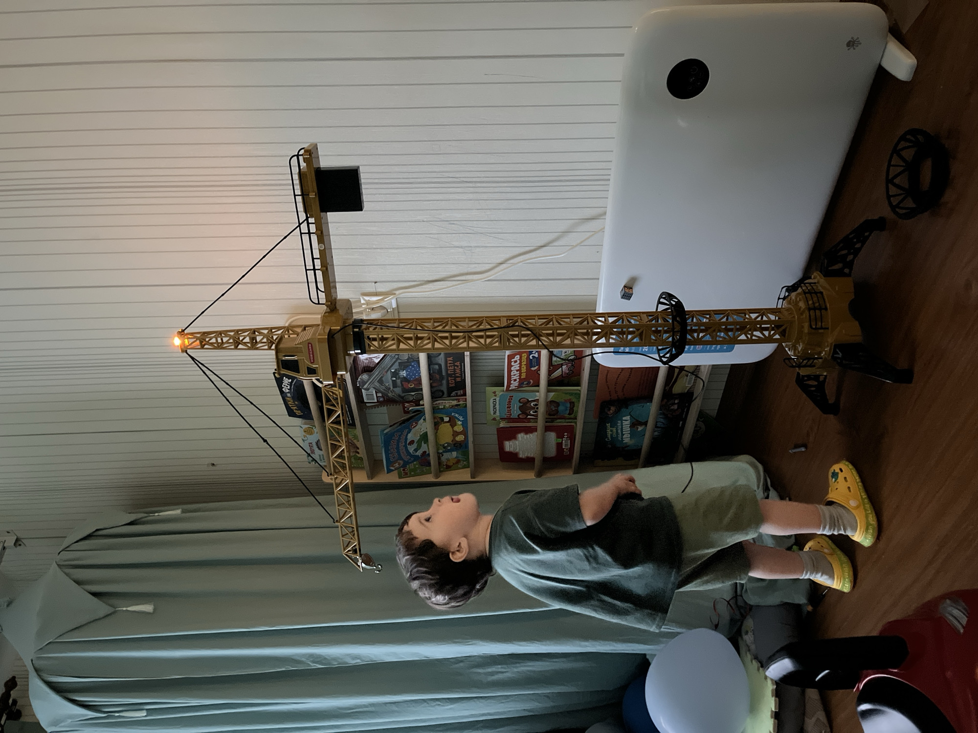 A child is standing in a room next to a toy crane and a bookcase filled with children's books written in Russian, including "ЗВЕРЯТА", "ПРЯТКИ ФЕРИЕ", "Сладких", "MDILIKA", and "КОТ, ПЕТУХ И ЛИСА". The child is wearing a green t-shirt, light green shorts, and yellow clogs. The toy crane has a working light on its boom. The scene is indoors, likely in a home.