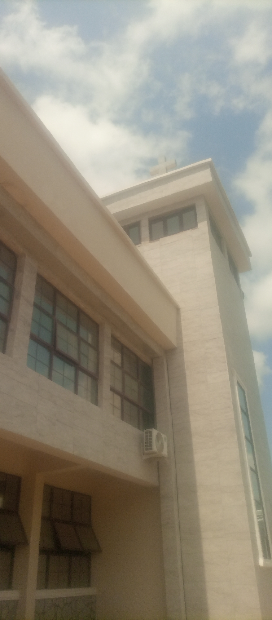 The image shows the exterior of a modern building with a light-colored, marble-like facade. The building features multiple windows with dark frames, some of which appear to be multi-paned. A beige overhang runs along a section of the building. An air conditioning unit is visible mounted on the exterior wall. The top of the building is visible against a blue sky with scattered white clouds. A cross-shaped structure is visible on the rooftop of the tallest section of the building, suggesting it might be a religious institution or have religious architectural elements. The scene is likely an outdoor view of a building in the city of Kuson, Nigeria. There are no people visible in the image.