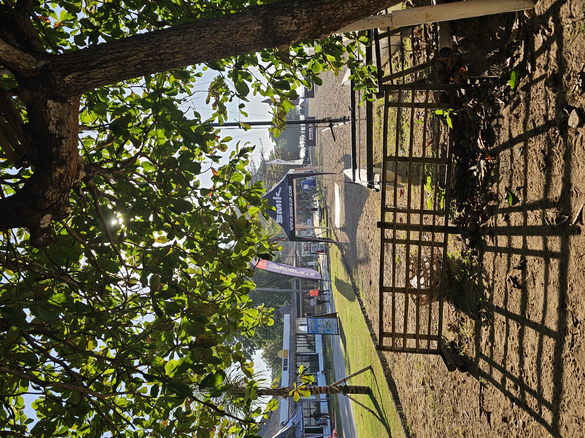 The image captures an outdoor scene in Denpasar, Indonesia, likely near a beach. A large tree with dense green foliage dominates the left side of the frame, its branches partially obscuring the view. Sunlight filters through the leaves, creating bright highlights.
On the right, a brown wooden fence stands next to a sandy area. The fence casts a distinct shadow on the ground. Beyond the fence, there's a grassy area and a building complex. A large promotional archway and various flags are visible further in the distance. People are present but appear small and are positioned along the path. The scene is brightly lit.