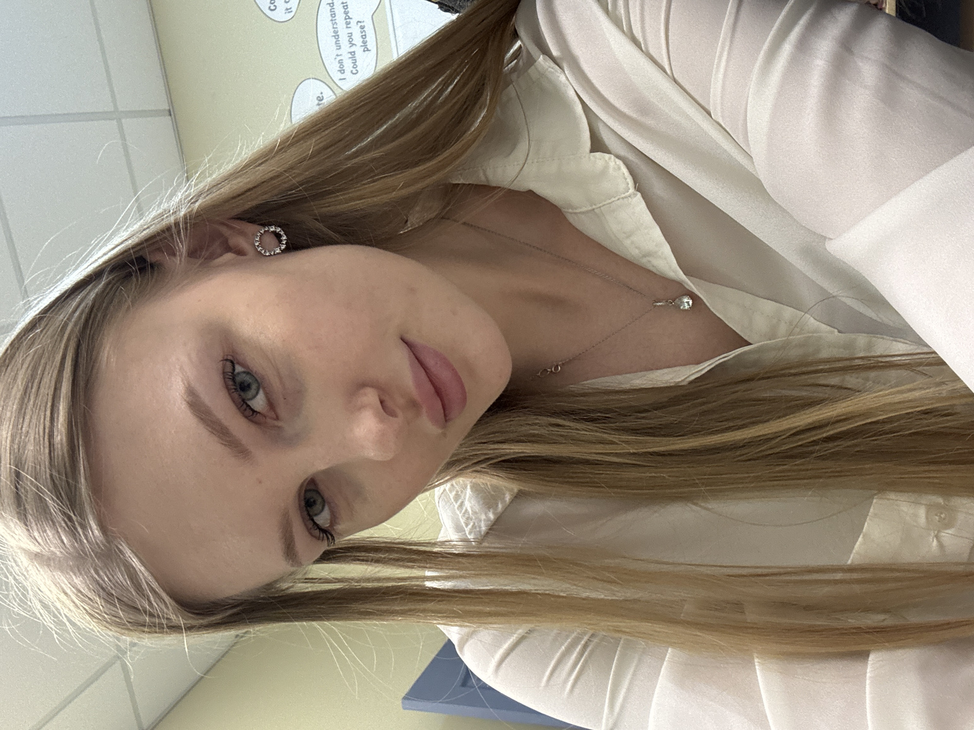 A fair-skinned female with light blonde hair is positioned in the foreground, wearing a white collared shirt and a delicate silver necklace. She has light blue eyes and is wearing a stud earring in her left ear. The background consists of a light green wall with a section of a sign containing the words "I don't understand. Could you repeat please?" visible in the upper left corner. The overall environment appears to be indoors, potentially a classroom or office. There is no indication that this image was taken in Perm, Russia.
