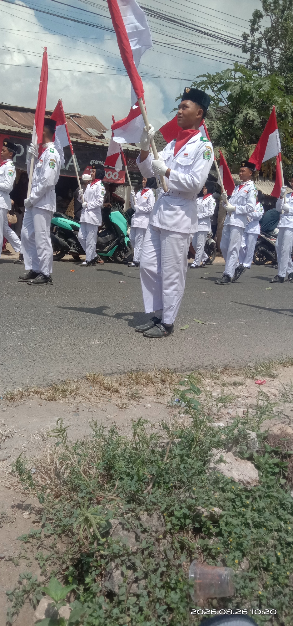 The image depicts a group of individuals in uniform marching in what appears to be a parade on a street in Pakong, Indonesia. The participants are wearing white uniforms with red neck scarves and black caps, and are holding Indonesian flags (red and white). They are walking on a paved road, with some vegetation and buildings visible in the background. A teal scooter is parked on the left side of the road, and another scooter is partially visible on the right. The sky is clear and bright, suggesting daytime. The overall scene conveys a sense of organized activity and national pride.