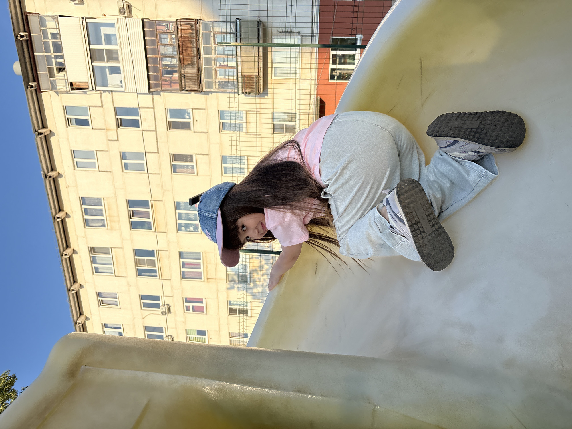 A young girl with long, dark hair and a blue and pink baseball cap is climbing up a large, cream-colored plastic slide on a sunny day. She is wearing a light pink t-shirt and light grey pants, and grey sneakers. The slide is part of a playground. In the background, a multi-story apartment building with a light yellow facade is visible. The building has many windows and balconies. A wire fence is in front of the building. The sky is clear and blue. The image appears to have been taken in Chaikovsky, Russia.