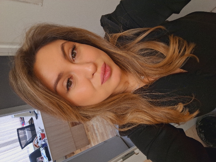 A forensic examination of the image reveals a close-up, chest-up portrait of a single adult female in an indoor setting. The subject, facing forward and looking directly at the camera, has fair skin, long wavy light-brown hair with lighter highlights, brown eyes with visible dark eyeliner and possibly false eyelashes, and full lips coated with pink gloss. She wears a black v-neck garment and a silver-colored necklace with a small pendant. Her expression is neutral with a slight subtle smirk or pout.
The background, partially visible and slightly out of focus, indicates an office environment. To the left of the subject, a light-colored wooden desk is seen with a black mesh office chair, a computer monitor, and various desk items, including a red and white polka-dotted cup. A window with vertical blinds is present further left. The floor appears to be light-colored wood or laminate. A dark-colored, possibly trash, container is partially visible in the lower left foreground. A light-colored wall forms the right side of the background.
There is no visual information within the image to determine the specific city as Khmel'niki, Russia, or any particular geographical location. The scene is consistent with a modern office interior.