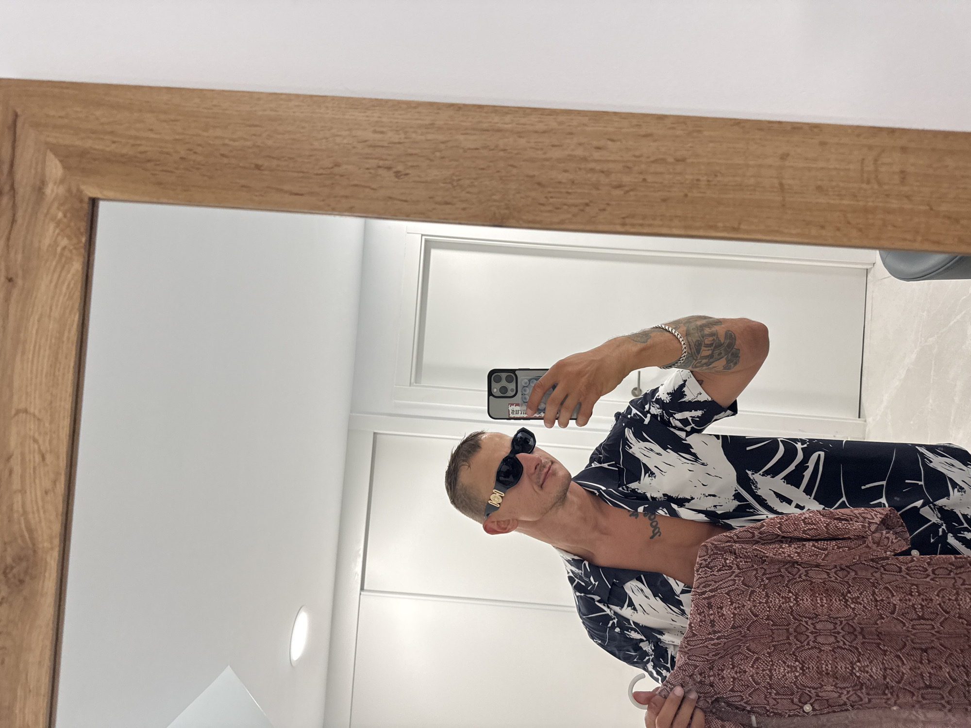 A man with short, dark hair and tattoos on his chest and arms is reflected in a large, rectangular mirror with a wooden frame. He is wearing black sunglasses with a gold emblem and an open, black and white patterned shirt. In his left hand, he holds up a pink, snake-skin patterned shirt. He is holding a smartphone in his right hand, as if taking a selfie in what appears to be a bedroom or dressing area. The background is a clean, white room with white sliding doors. The floor is tiled with a light grey pattern. This image was taken in Finestrat, Spain.