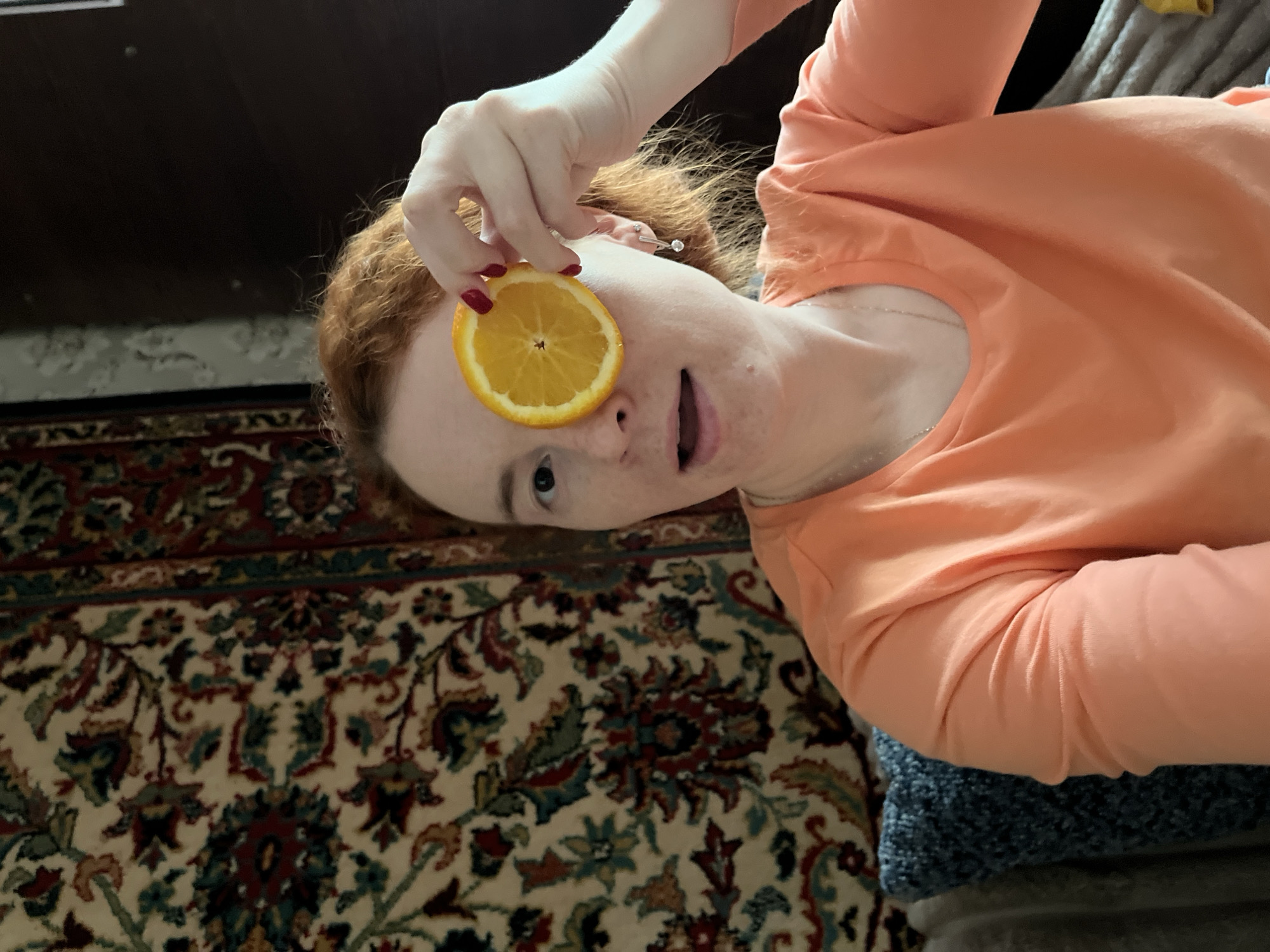 A woman with red hair and freckles is holding a slice of orange over one eye. She is wearing an orange long-sleeved shirt and a silver necklace. Her fingernails are painted red. She is looking directly at the camera with a surprised expression. The background includes a patterned rug and a dark wooden piece of furniture. The image was likely taken indoors. It is not possible to confirm the location as Chaikovsky, Russia, without additional information or evidence.