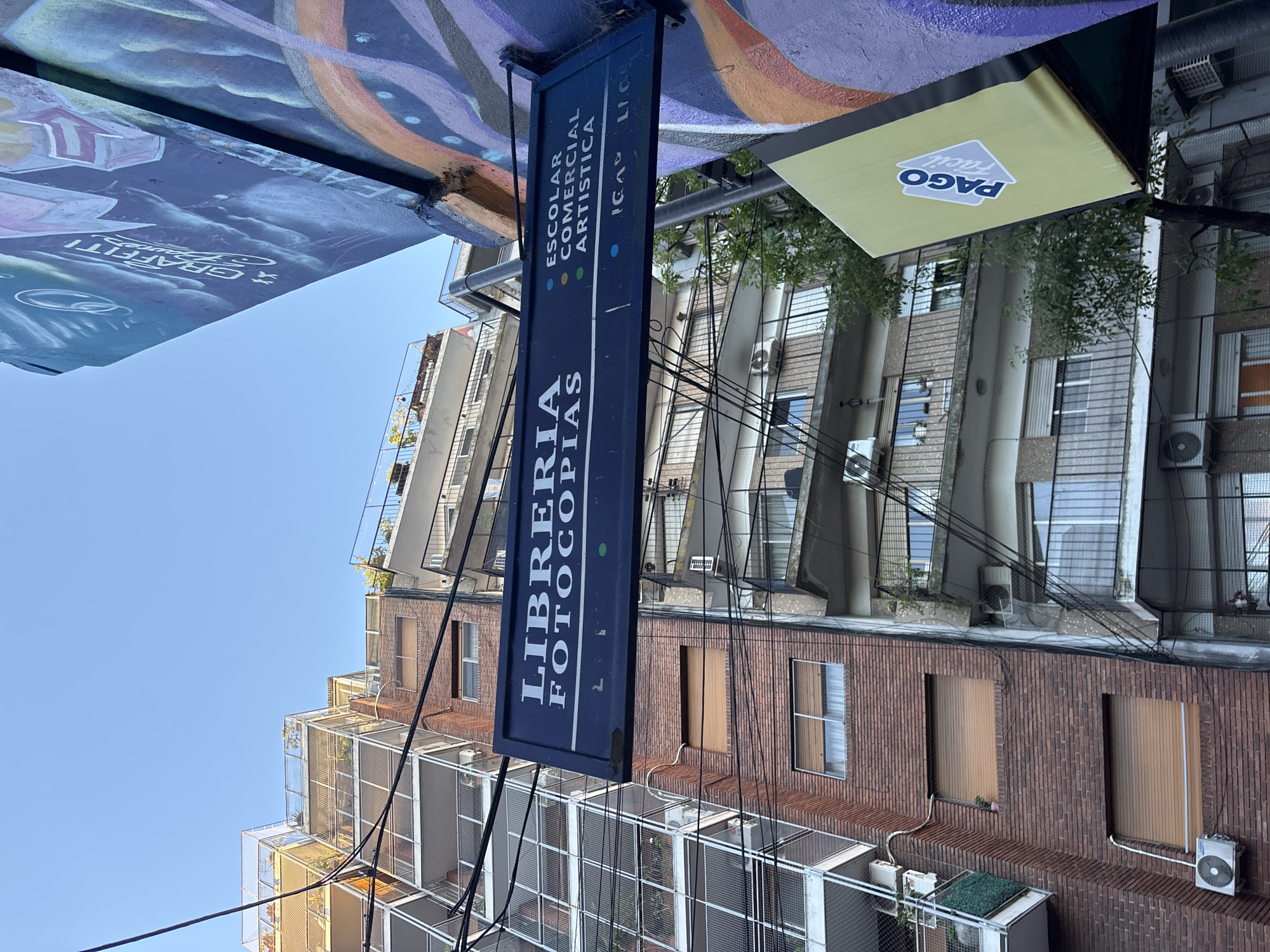 The image depicts a street scene in Buenos Aires, Argentina. A dark blue sign with white lettering is prominently displayed, reading "Libreria Fotocopias" and "Escolar Comercial Artistica ICAV LICS." The sign hangs beneath a colorful mural. Buildings with a mix of brick and concrete facades, some with screened balconies, are visible in the background. A yellow sign with "Pago Facil" is also visible. Many utility wires crisscross the sky. The sky is clear and blue.