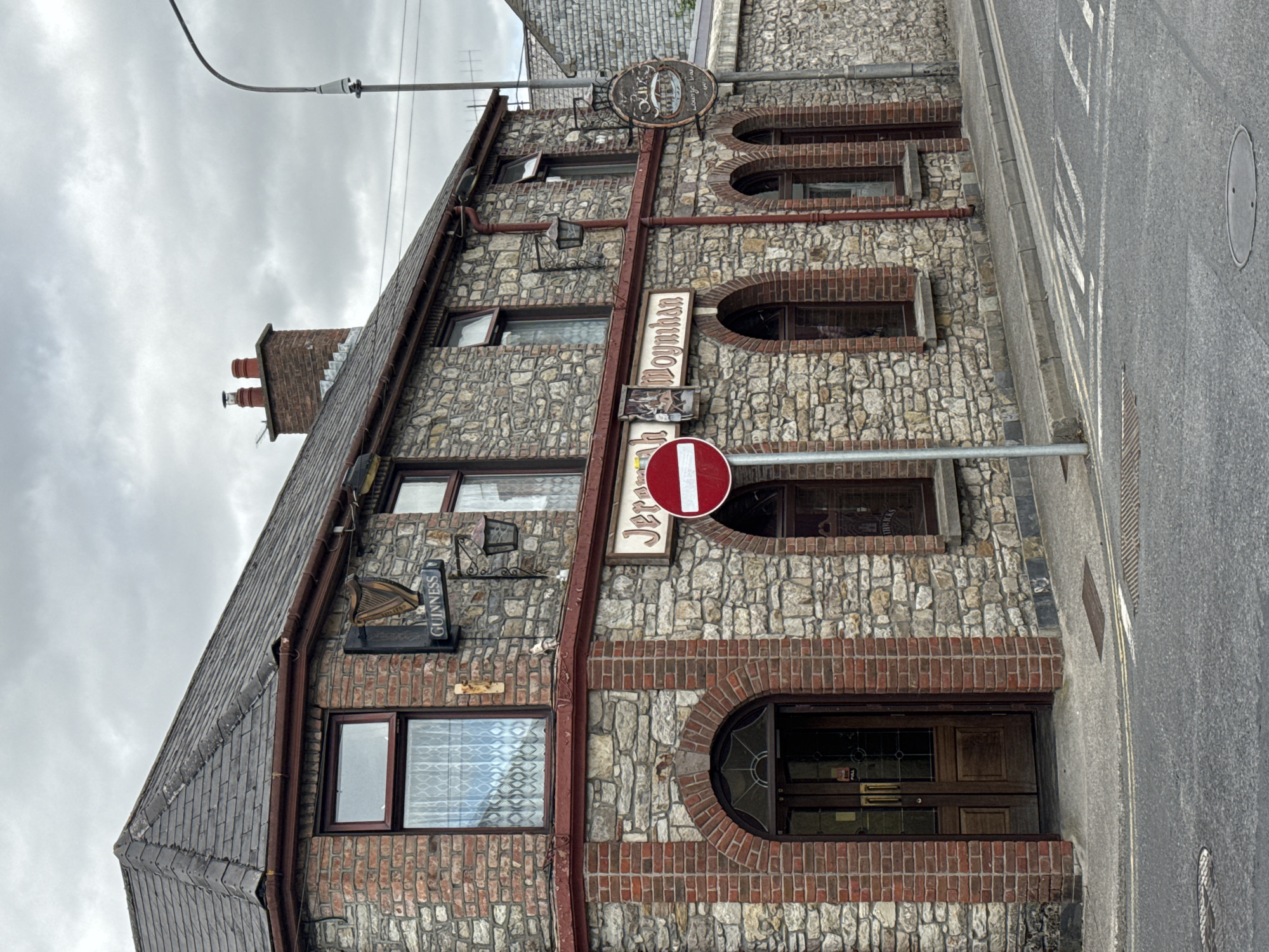 This is a daytime, eye-level, exterior, medium shot of Jeremy Moynihan's Pub, located on a street in Clonmel, Ireland. The pub is a two-story building with a stone facade and red brick accents around the windows and entrance. The roof is slate. A Guinness sign is visible on the upper left of the building, and a circular sign that appears to say "O'Malley's Lounge Bar" is on the right. A red and white "no entry" sign is posted on a pole in front of the pub. The street in front of the pub has double yellow lines painted on it, indicating a no-parking zone, and a pedestrian crossing. The sky is overcast. There are no people visible in the image.