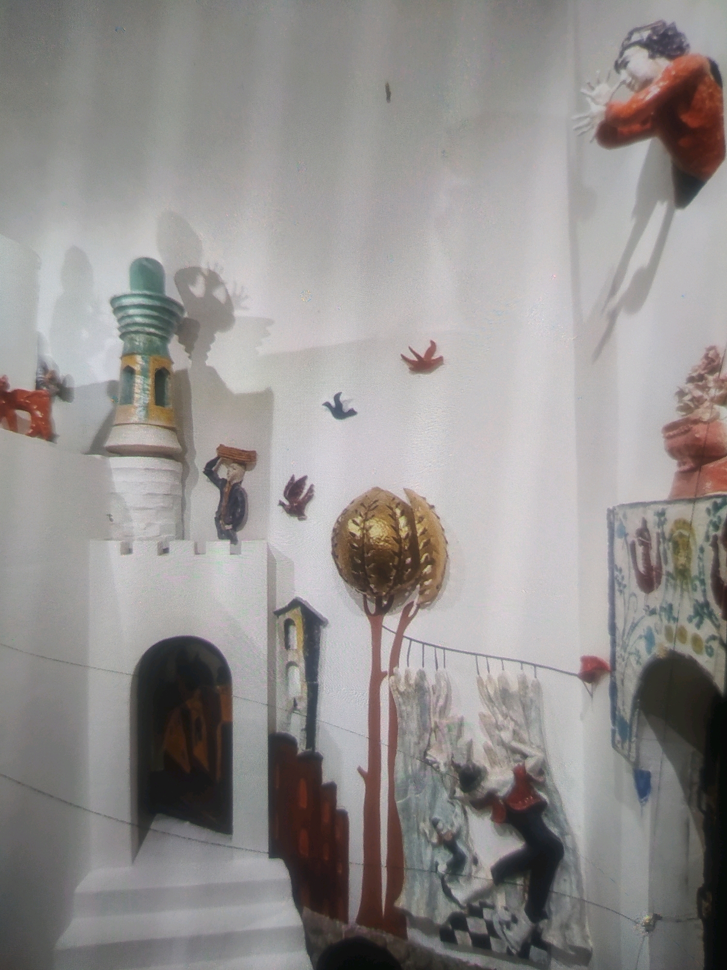 The image shows an installation art piece displayed on a white wall. The installation features several sculpted figures and elements arranged to create a surreal, dreamlike scene.
On the upper right, a figure in a red shirt and black pants is depicted emerging from the wall, with hands outstretched as if trying to push off. This figure casts a long shadow.
To the left of this figure, a small, stylized red bird-like shape is mounted on the wall. Further to the left, a larger, detailed red bird sculpture is also visible.
In the center of the image, a tall, white, castle-like structure is prominent. It has a rounded archway at its base, revealing a dark interior. Atop this structure, a figure in a dark outfit is shown carrying a rectangular object on its head, facing left. Above this figure, a small blue bird is mounted on the wall.
To the left of the castle structure, a red, animal-like sculpture is partially visible. A green and yellow tower-like sculpture stands next to the castle.
In the lower central part of the image, a golden, bulbous object resembling a seedpod or a fantastical tree head is suspended by thin wires, attached to a stylized brown tree trunk. A black and white checkered pattern is visible on the wall below this object.
To the right of the checkered pattern, a relief sculpture depicts two figures. The larger figure, dressed in red and black, is in a dynamic pose, seemingly falling or dancing on the checkered surface. A smaller figure is also visible behind and below it.
Further to the right, a section of a decorative tiled structure with floral motifs is visible, with a terracotta-colored potted plant or sculpture on top.
The overall environment appears to be an indoor exhibition space with plain white walls serving as the backdrop for the art installation. There are no indications of the specific location within the city of Birlik, Uzbekistan.