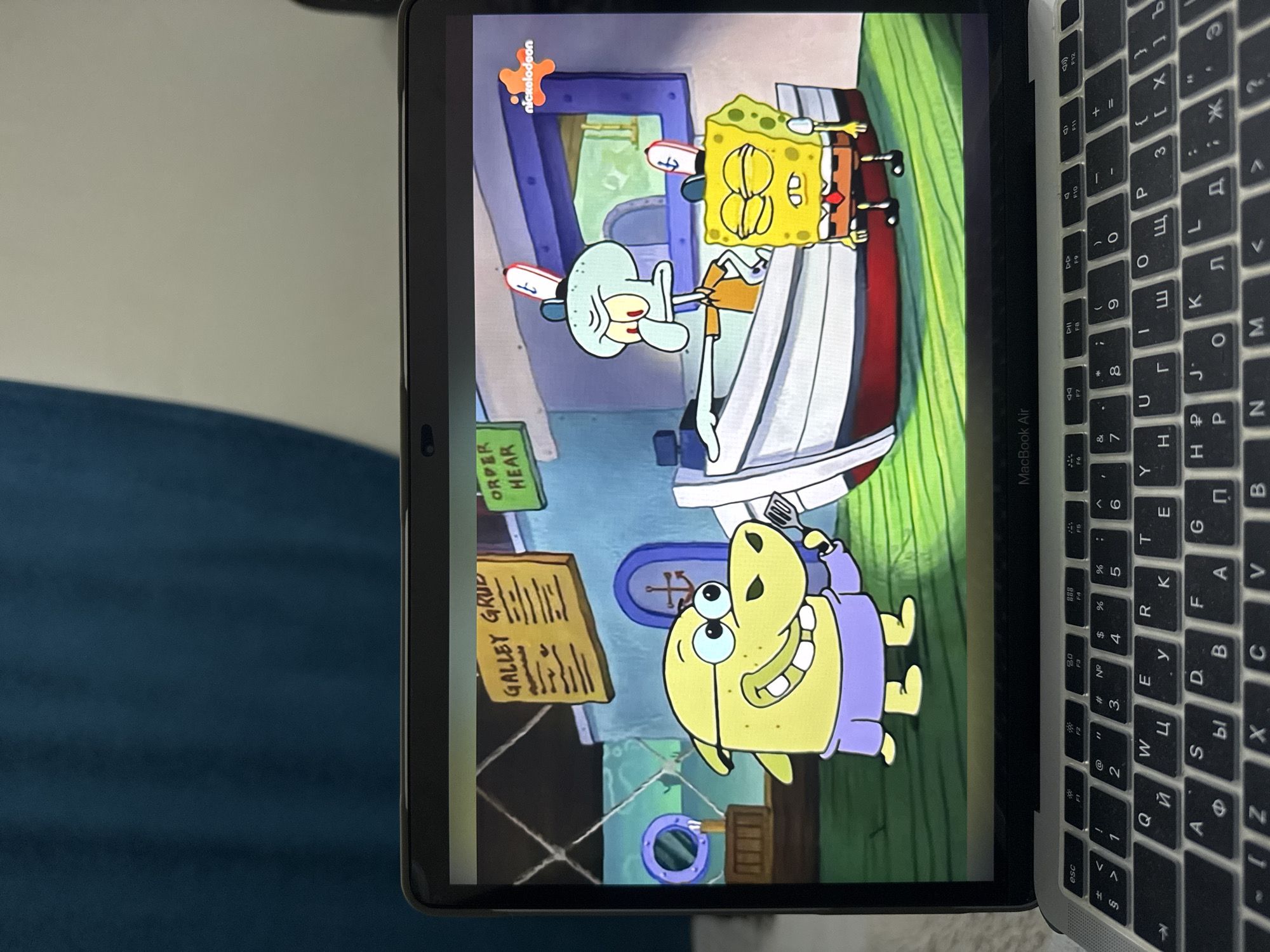The image shows a laptop displaying a scene from the animated television show SpongeBob SquarePants. On the screen, three characters are visible: a yellow hippopotamus-like character wearing glasses and a purple shirt, holding a spatula; Squidward Tentacles, a light blue octopus-like character with a stern expression, gesturing with his left hand; and SpongeBob SquarePants, a yellow sea sponge character with closed eyes and a tired expression, wearing a white shirt, red tie, and brown pants. They are positioned in front of a boat in what appears to be an outdoor setting with a blue and white building in the background. A sign labeled "GALLEY GRUD" and another green sign are also visible on the building. A Nickelodeon logo is present in the upper right corner of the screen. The laptop itself is open and placed on a surface. The background behind the laptop shows a dark blue fabric. There is no indication or visual evidence within the image that places this scene or the laptop in the city of Şişli, Turkey.