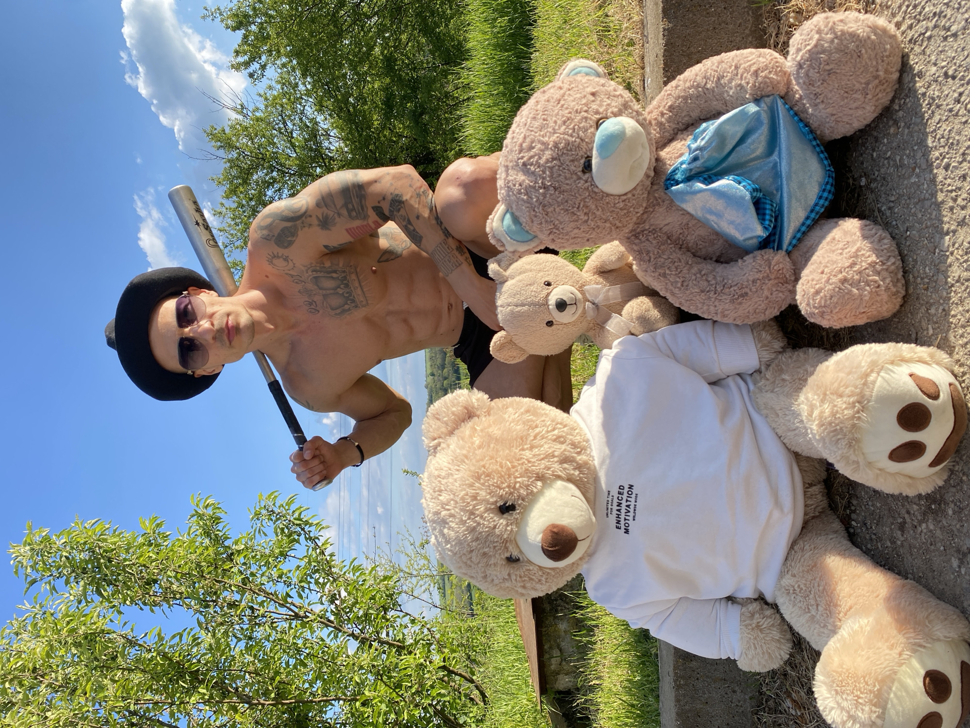 A low-angle shot shows a muscular, shirtless man wearing sunglasses and a black hat, standing next to three stuffed teddy bears on a concrete surface outdoors on a sunny day. The man is holding a baseball bat and has numerous tattoos on his upper body. The teddy bears are light brown; one is wearing a white t-shirt with text on it, another has a blue square of fabric on its stomach, and the smallest is sitting in the middle with a white ribbon on its neck. Green trees are in the background and the sky is blue with scattered white clouds. The location appears to be in the city of Pleven, Bulgaria.