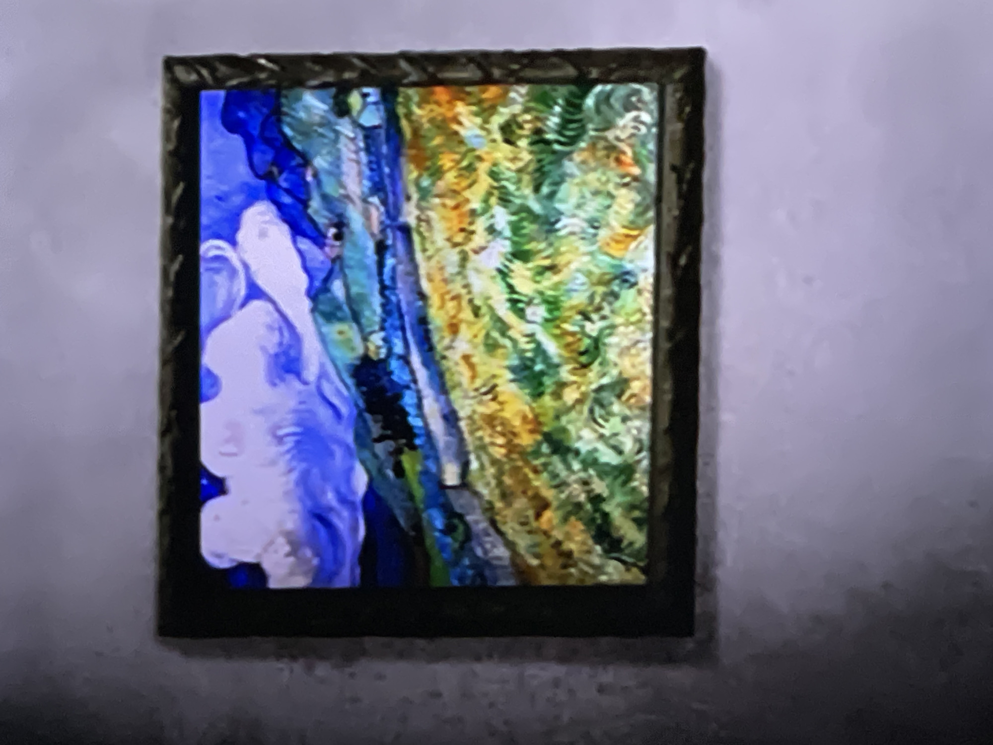 A framed abstract artwork is displayed on a light-colored, possibly off-white or light lavender, wall in an indoor setting. The frame is dark, appearing to be made of wood, and features intricate, possibly carved, decorative details.
The artwork itself is vibrant and highly textured. It is predominantly divided into two vertical sections. The left section is dominated by shades of blue and white, with fluid, organic shapes that suggest a swirling, cloud-like, or water-like abstraction. The right section features a rich tapestry of greens, yellows, and oranges, with a distinct, almost impasto-like brushwork that creates a textured, foliage-like or landscape-inspired pattern. A narrow vertical strip, featuring shades of blue and grey, separates these two dominant color fields.
No specific activity or event is taking place; the image captures a static display of art. The time of day and weather conditions cannot be determined from the indoor setting. There are no identifiable location cues, landmarks, or visible text within the image that confirm a specific location like Moscow, Russia. The overall image quality is somewhat soft and slightly noisy.