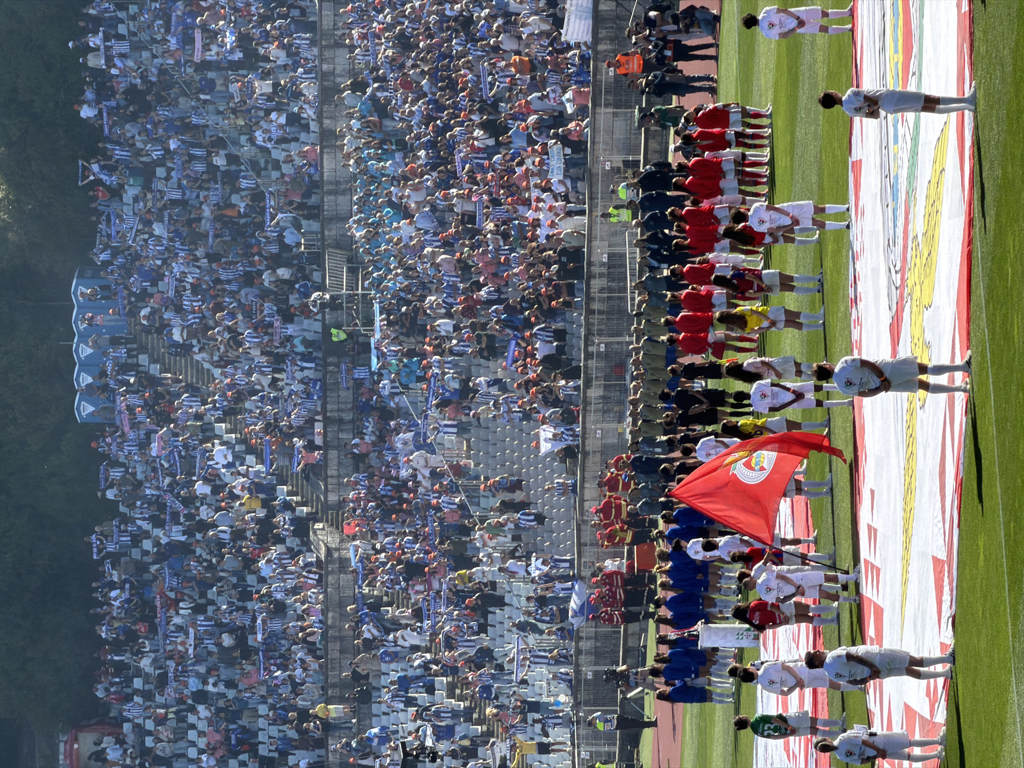 The image shows a soccer match taking place in Oeiras, Portugal. A large crowd fills the stadium stands, waving blue and white scarves. On the field, two teams of players line up, with one team in blue uniforms and the other in red. A large red flag is being held near the front of the red team. A banner with red, white, and yellow patterns is spread on the field in front of the teams. The scene is bathed in sunlight.