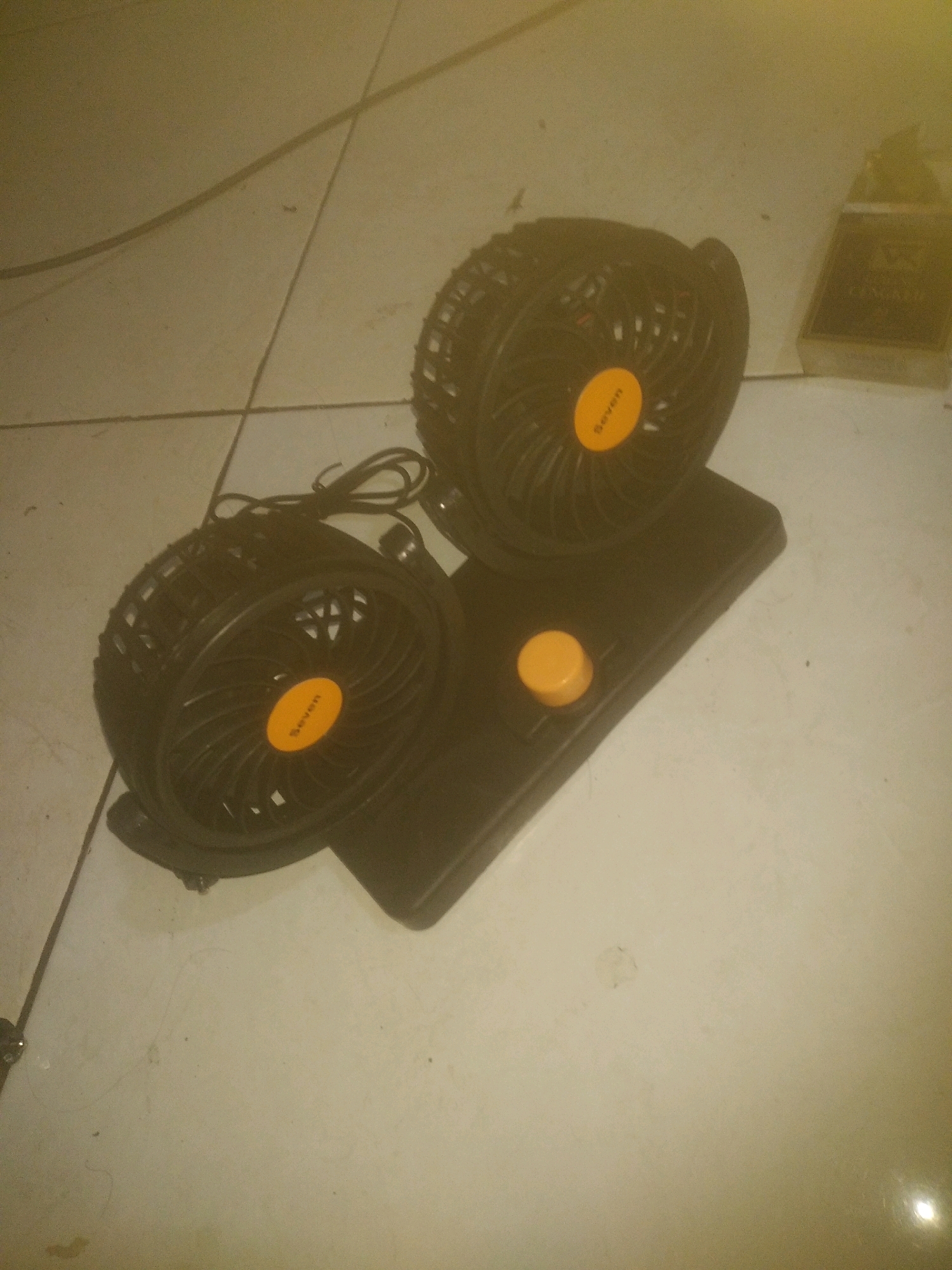 The image shows a black dual-fan portable device placed on a tiled floor. The device has two black fans with orange circular logos that read "Seven". A single orange button is visible on the base of the device. To the right of the device, a portion of a cigarette pack is visible. The lighting in the image is dim, casting shadows and giving the scene a yellowish hue. The floor is tiled with visible grout lines. The environment appears to be indoors, but the specific location within a city cannot be determined.