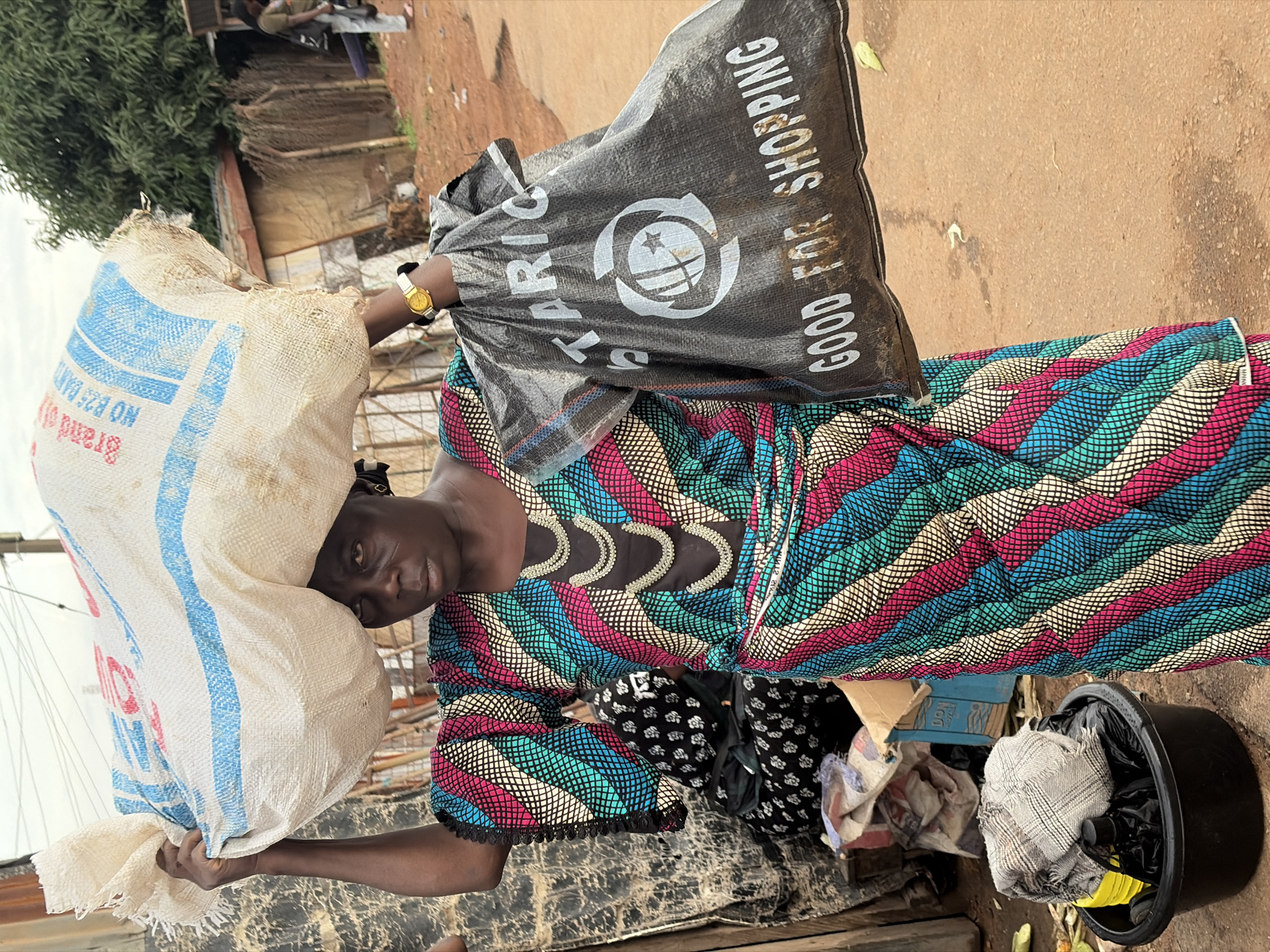 A Black woman in Gura Top, Nigeria, carries a large, white, woven sack of goods on her head, her left arm extended upwards to support it. She is also holding a black reusable shopping bag in her right hand. The woman wears a brightly colored dashiki-style dress with a geometric pattern of pink, turquoise, white, and black. She has a gold watch on her left wrist. In the background, there are wooden structures, a large green tree, and other people. The ground is reddish-brown dirt. There is a black bucket containing various items, including a checkered cloth and black plastic bags, to the woman's left. The overall scene depicts a street market environment.