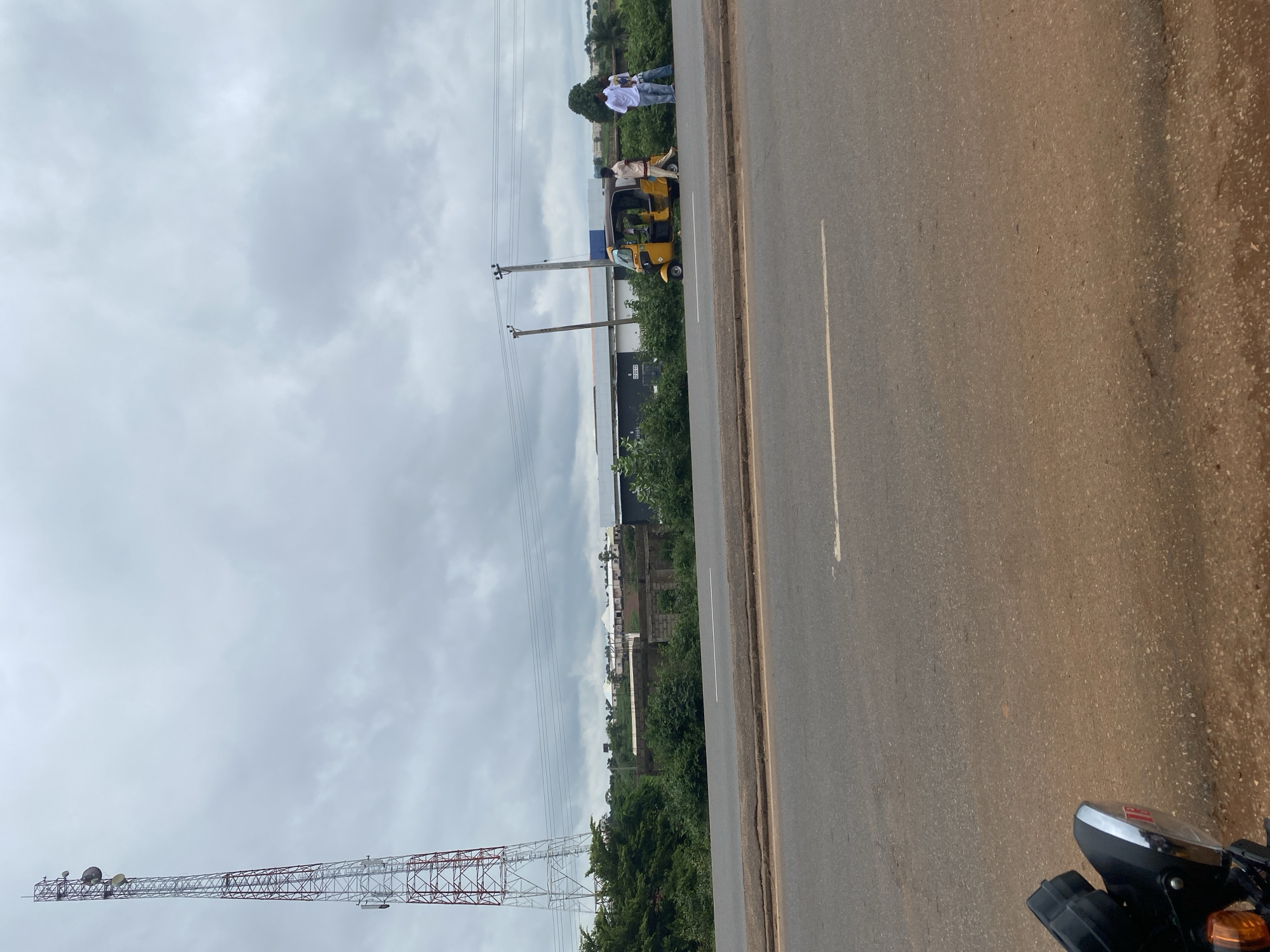 The image depicts a roadside scene in Lafia, Nigeria, under a cloudy sky. In the background, a tall, red and white telecommunications tower stands prominently on the left, with several antennas attached. Adjacent to the tower, a line of trees and lush green vegetation borders a low-rise building with a white facade and a dark, possibly corrugated metal, roof. Further along the road, to the right, a yellow auto-rickshaw (a three-wheeled vehicle) is parked. Two men are standing near the auto-rickshaw, and another man is walking away from it. They are all dressed in casual clothing. The road in the foreground is paved and shows signs of wear, with a white line marking the lane. A portion of a motorcycle's front is visible in the lower left corner of the image. The overall environment suggests an urban or semi-urban setting in Nigeria.
