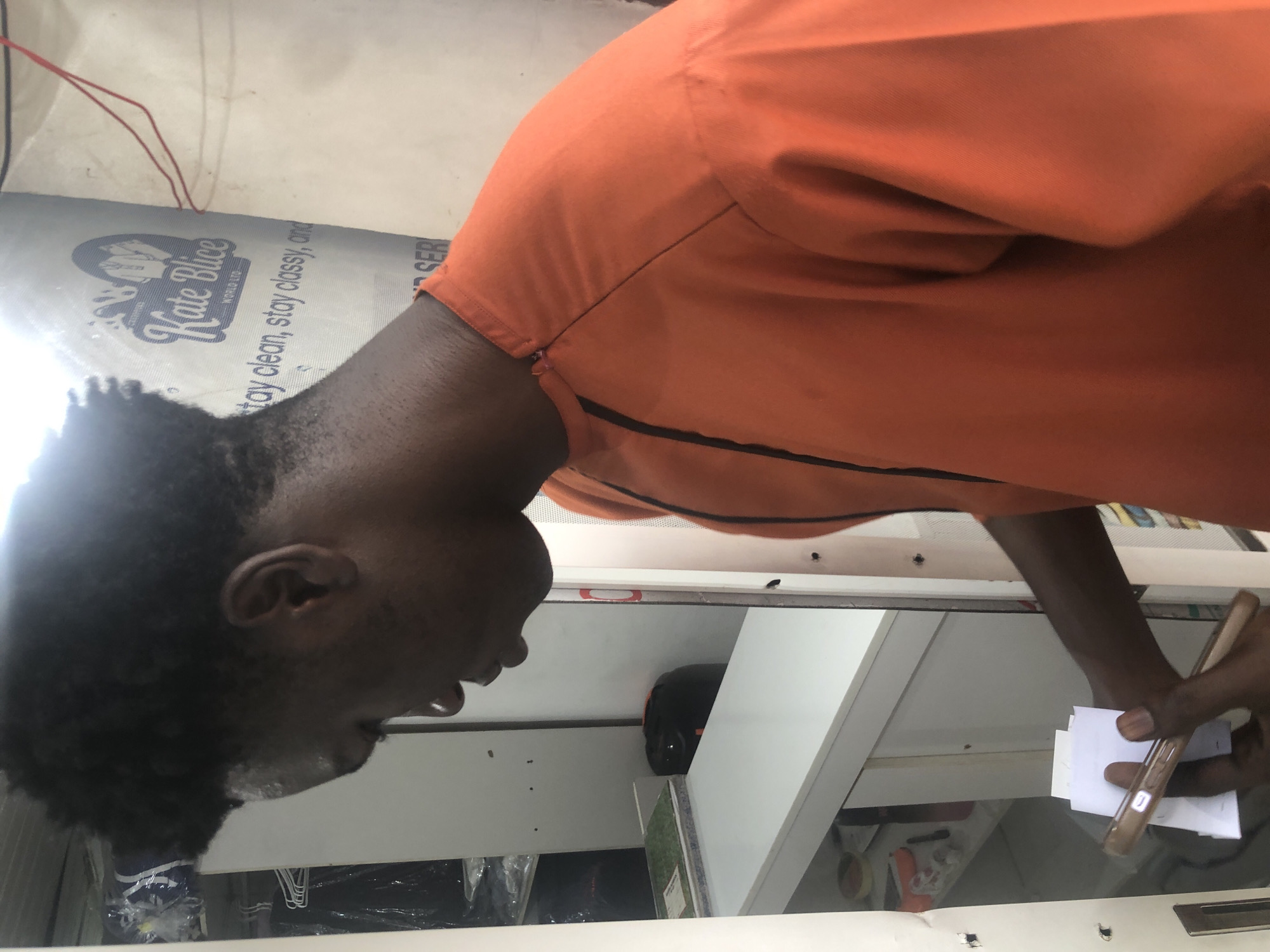A young Black man wearing an orange shirt is standing in front of a white counter with shelves behind it. He is holding a stack of white papers in his right hand and a phone in his left. A logo for "Kate Blice World Ltd." is visible on a blue and white banner in the background, with the slogan "stay clean, stay classy, and". The environment appears to be a service establishment, possibly a dry cleaner.