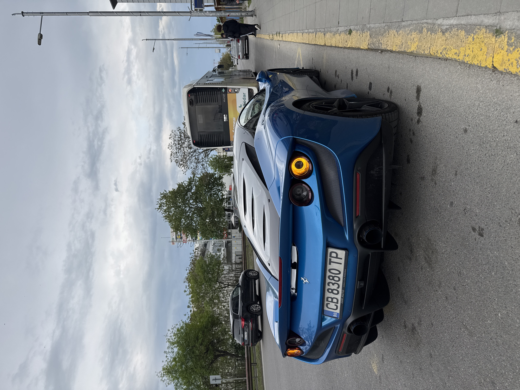 A blue Ferrari is parked on a city street in Plovdiv, Bulgaria. The license plate reads "CB 8380 TP." A bus is stopped in the adjacent lane. A person is walking on the sidewalk. The sky is cloudy. Trees and streetlights line the background. Other vehicles are parked further down the street. Yellow curb lines are visible.