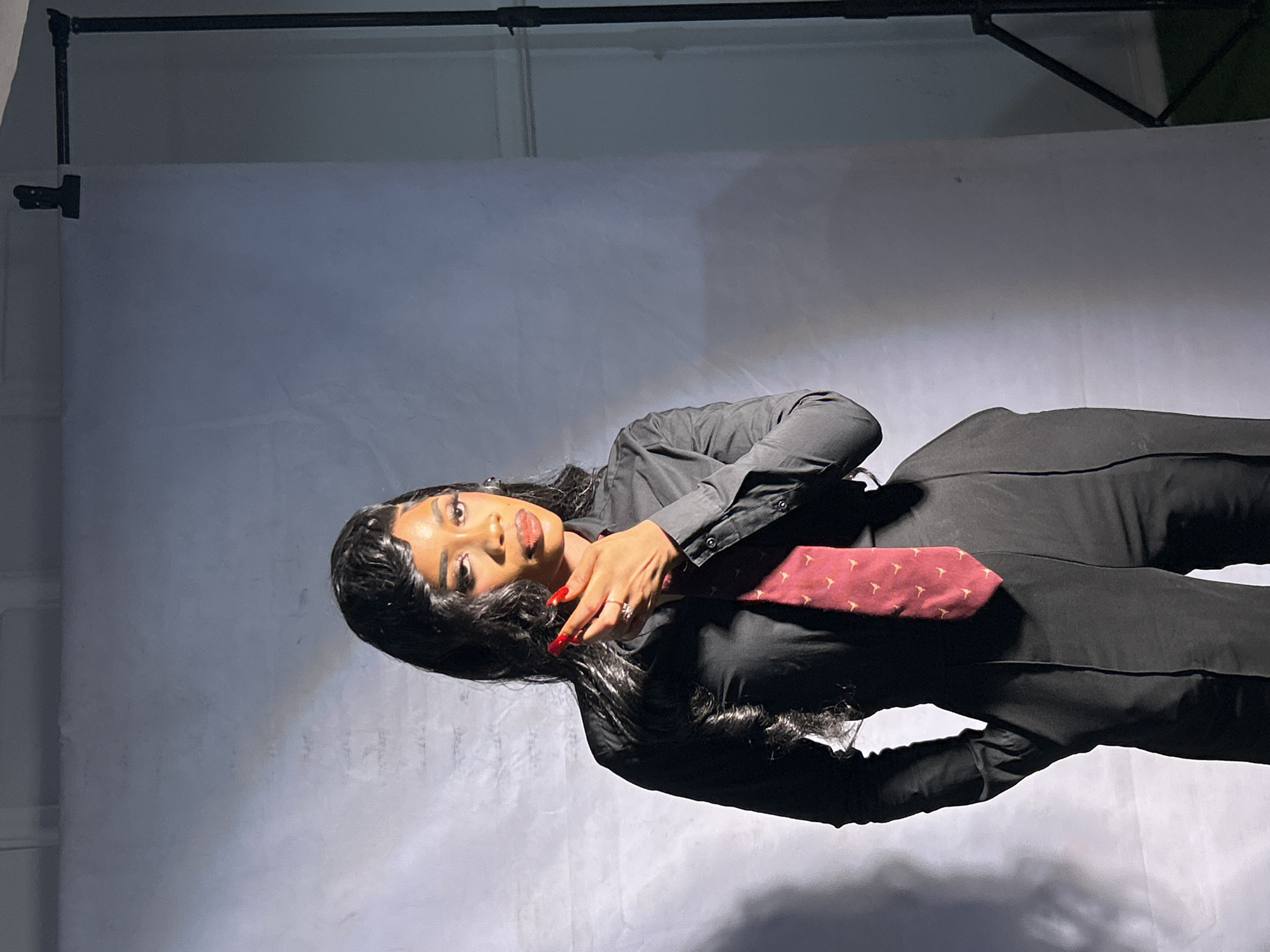 A young woman with long black hair and red nail polish, wearing a black button-down shirt, black pants, and a burgundy tie, is lying on a light grey backdrop. The backdrop is supported by a black metal frame. The setting appears to be a studio environment. There is no evidence to confirm if the image was taken in Jos, Nigeria.