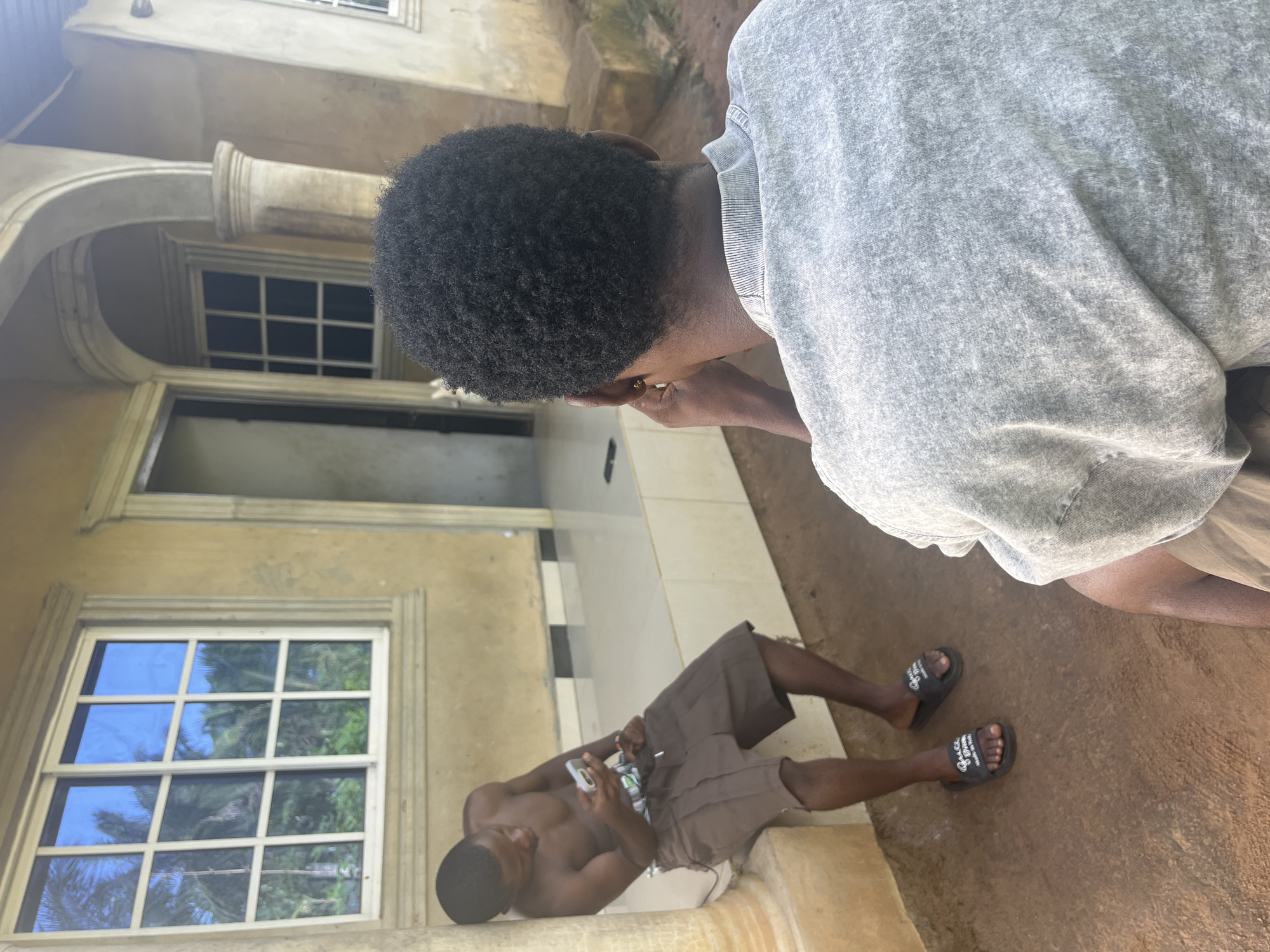 An eye-level shot depicting two Black men outside a two-story building. A man with short, dark hair and a gray sweatshirt is positioned to the right of the image, leaning forward and slightly out of focus. To the left, another shirtless man is lying down with brown shorts and black sandals; he is holding a device. The building has a cream-colored exterior with columns and windows with multiple panes. The reflections on the window suggest a tropical setting, likely somewhere in Ikot Ekpene, Nigeria. The surrounding ground is brown.
