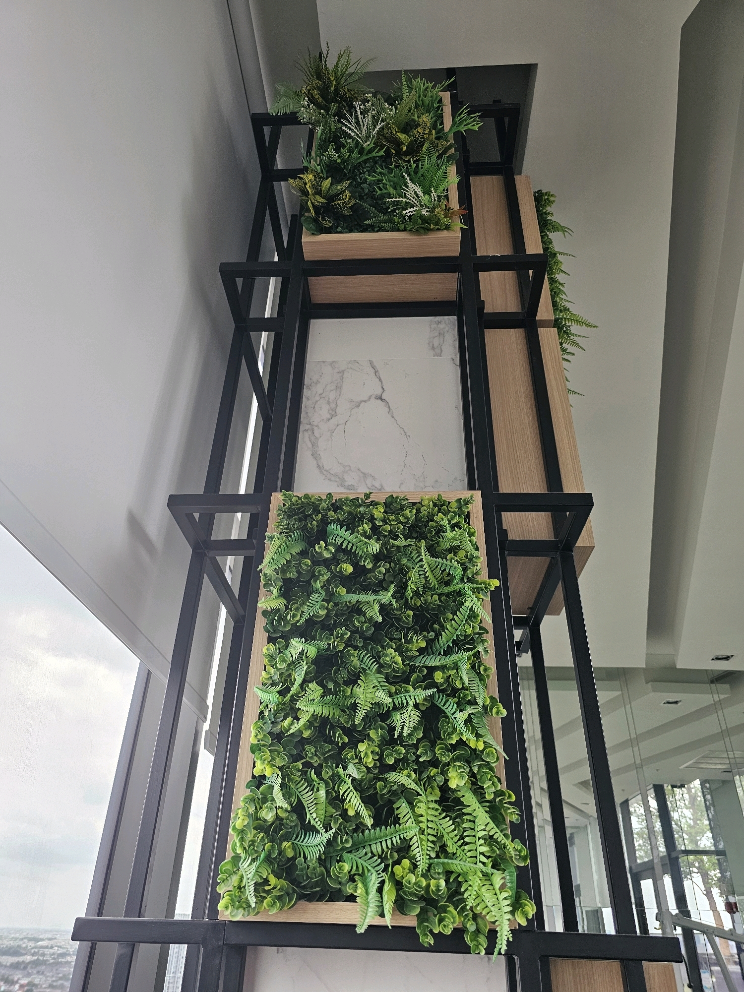 This is a low-angle, full shot of a tall, multi-tiered vertical plant display against a white wall and window, likely indoors at an office or public building in Bangkok, Thailand. The display is constructed with a black metal frame and light wood shelves. On the top shelf, a rectangular wooden planter holds an arrangement of various artificial green plants, including feathery ferns and leafy bushes. Below this, a shelf features a large, rectangular section filled with dense artificial greenery, mimicking a living wall. This bottom section is framed by a wooden border and showcases a variety of green leaves and ferns. In the background, the white wall transitions into a ceiling with modern architectural elements, and a large window reveals a cloudy sky and a distant cityscape, characteristic of Bangkok. There are no people or animals visible in the image. The overall impression is one of modern interior design incorporating artificial plant elements.