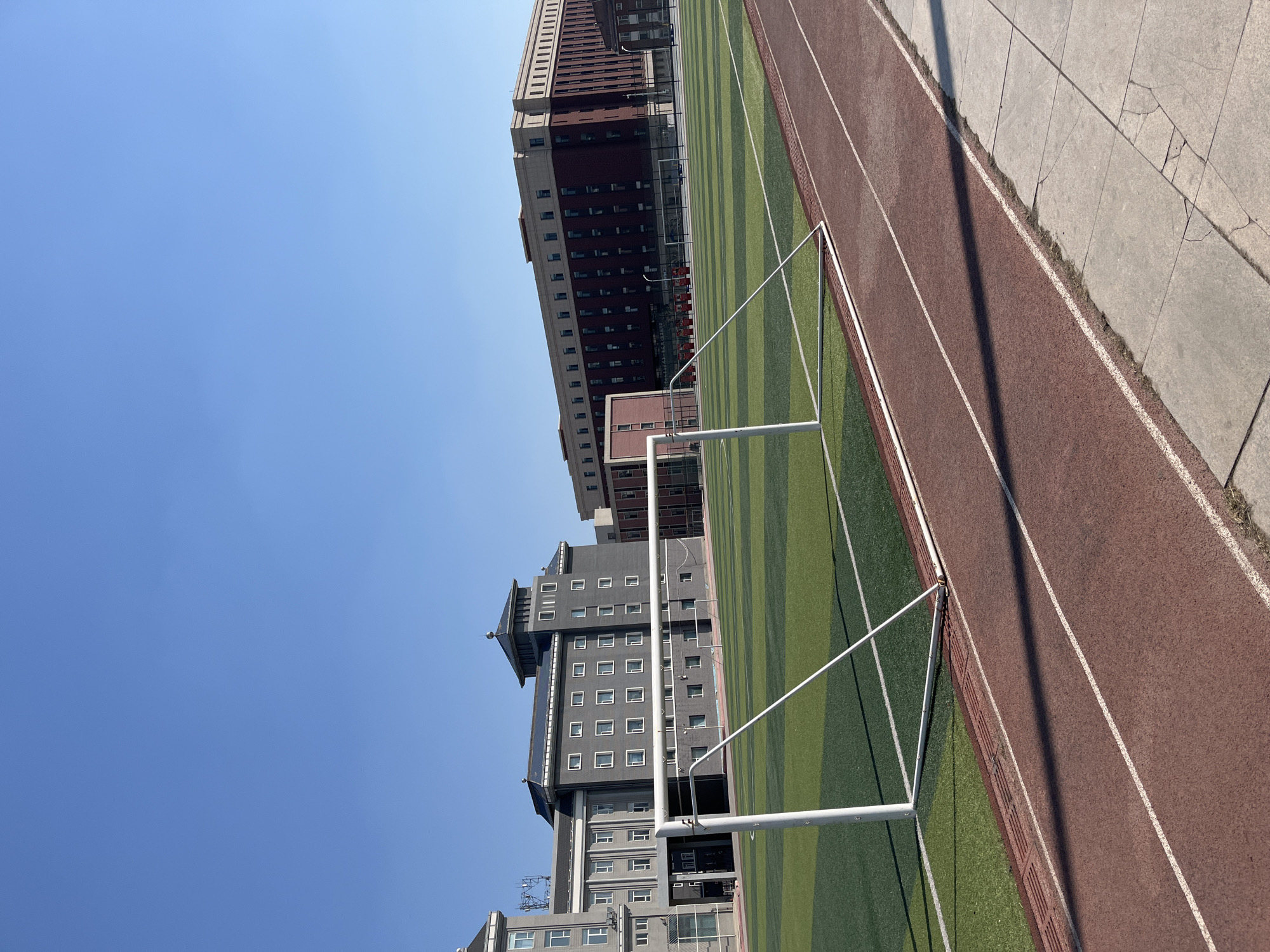 The image shows a sports field with a running track in Changchun City, China. A soccer goal stands on the green and darker green striped field. The running track is red. Adjacent to the track is a concrete area. Tall buildings are in the background. The sky is blue.