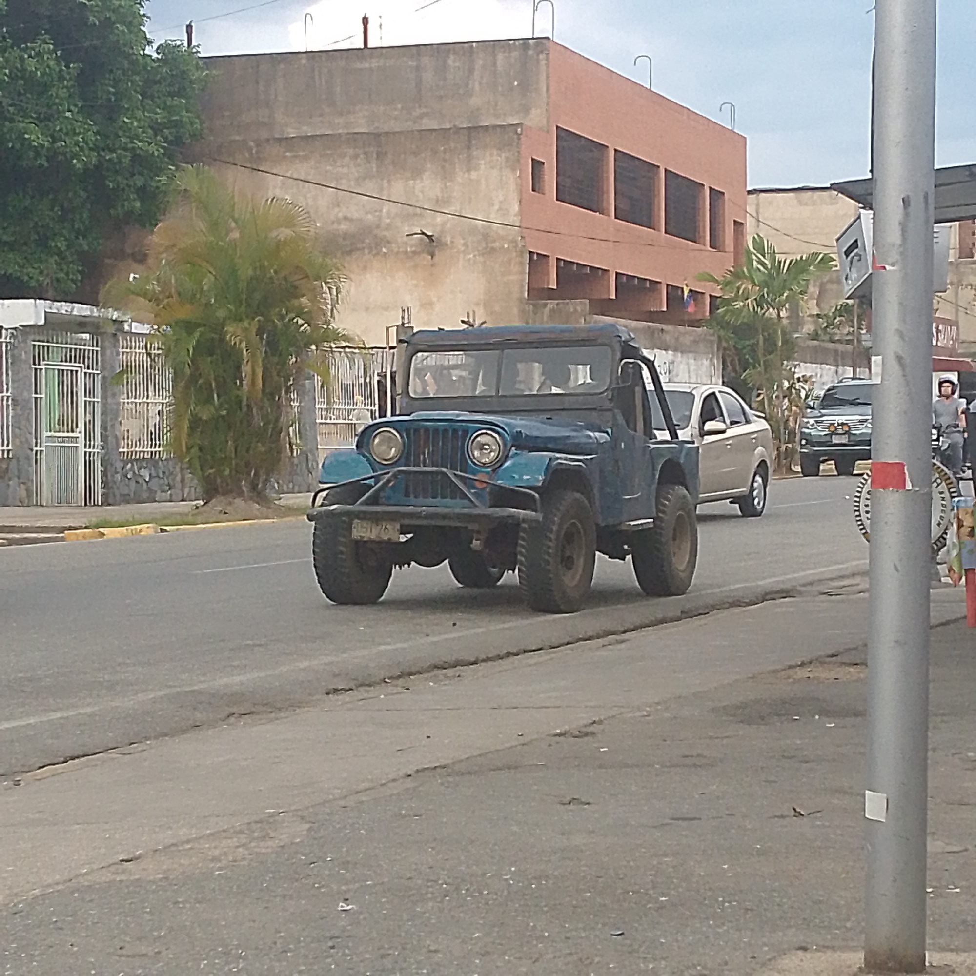 The image shows a blue Jeep-like vehicle driving on an asphalt road in Tinaquillo, Venezuela. Buildings of concrete construction, trees, and other vehicles, including a gold car and a dark SUV, are in the background. A light-colored metal pole with attached signage is in the foreground to the right. A person on a motorcycle wearing a helmet is visible to the right of the blue Jeep. The road surface is cracked and shows signs of wear. The sky is overcast.