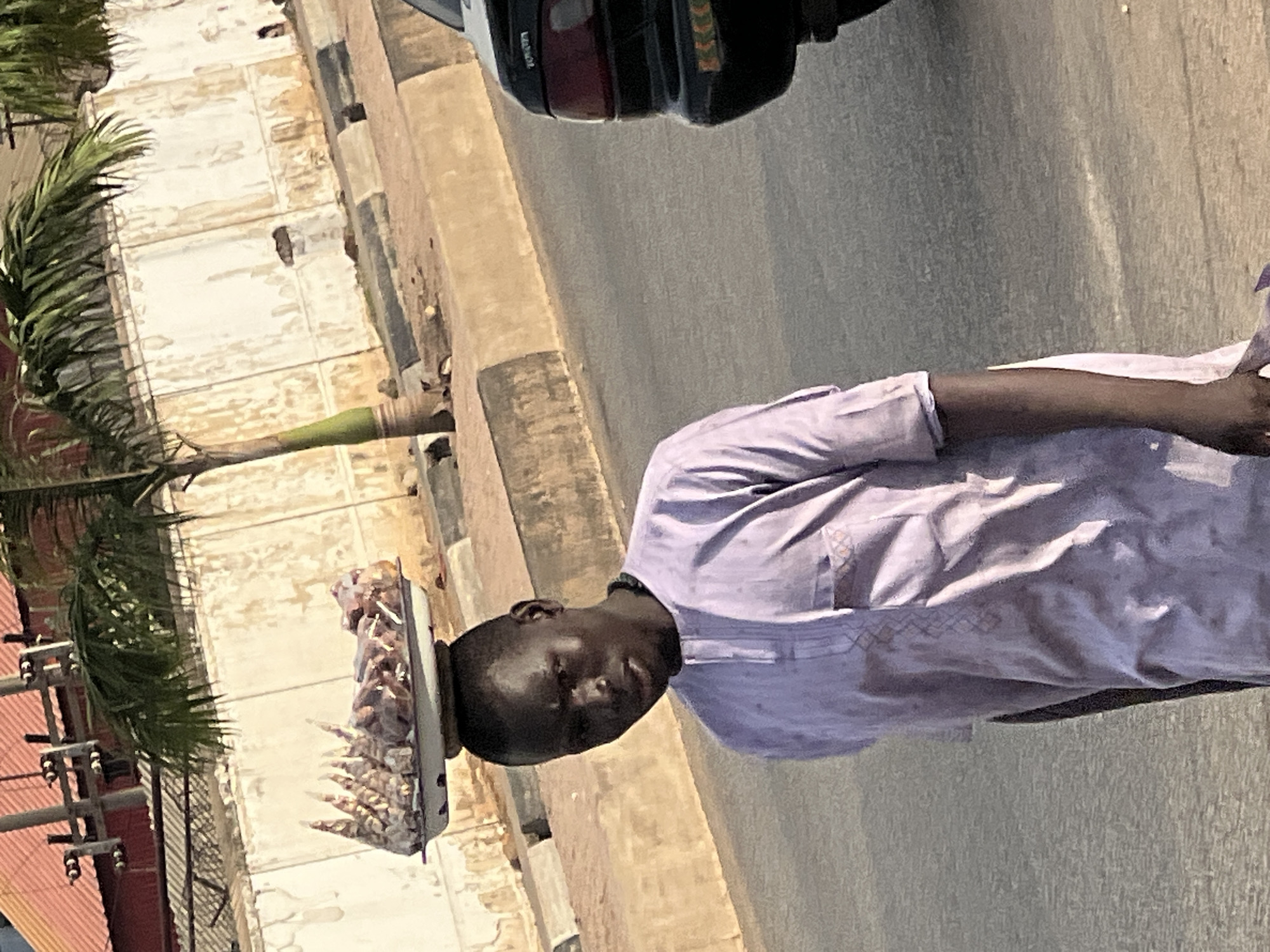 A man balances a tray of cooked meat on his head as he walks along a street in Barakin Lerawi, Nigeria. He is wearing a light purple, short-sleeved shirt. A vehicle is partially visible at the top of the frame. A palm tree and a building are in the background.