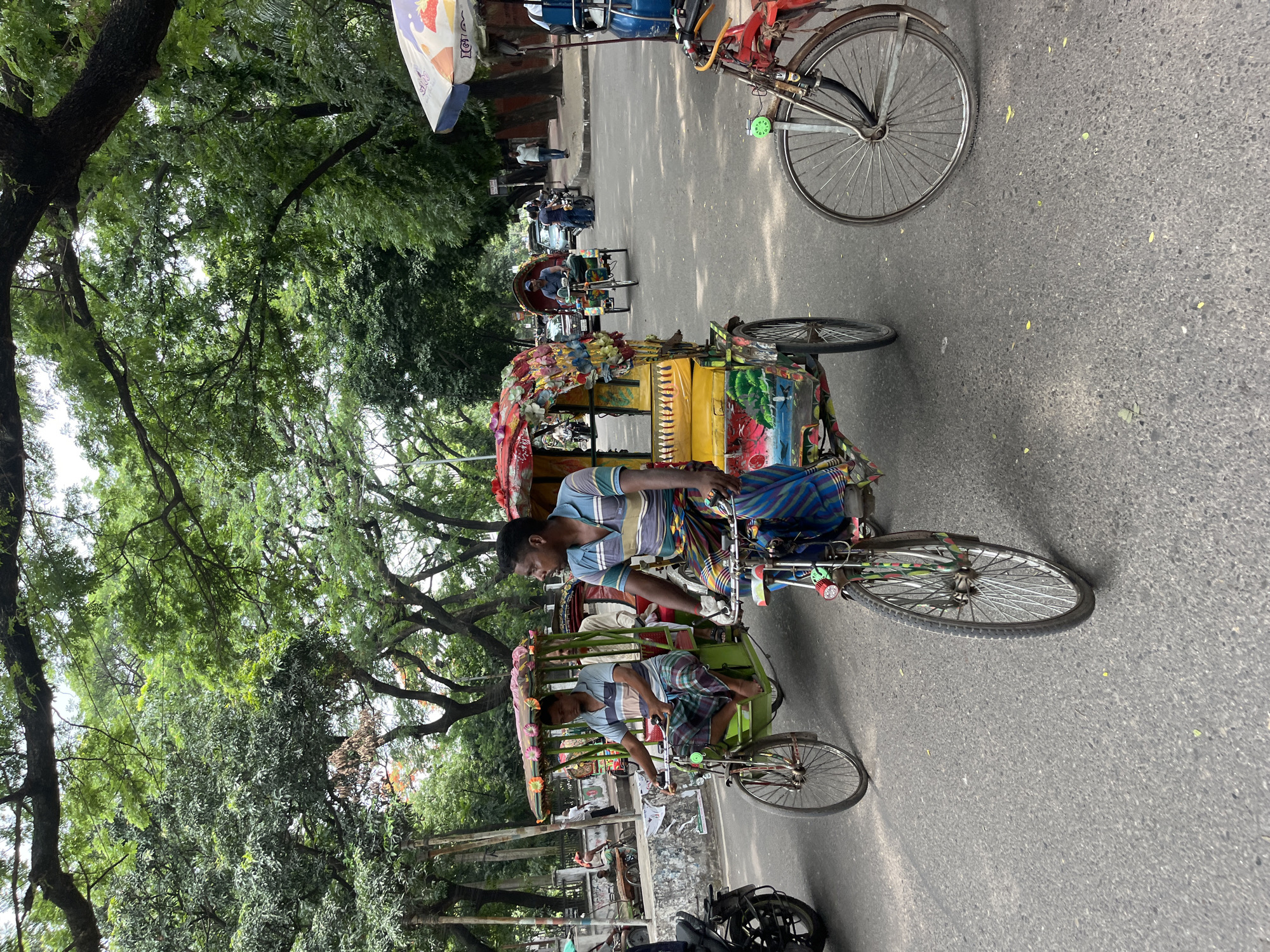 The image depicts a street in Dhaka, Bangladesh, featuring two rickshaws. The rickshaws are elaborately decorated with colorful paint and ornaments. The drivers are dressed in striped shirts and lungis, traditional garments. The street is paved and lined with trees. There are other rickshaws and pedestrians in the background. The environment suggests a busy urban setting in Dhaka.