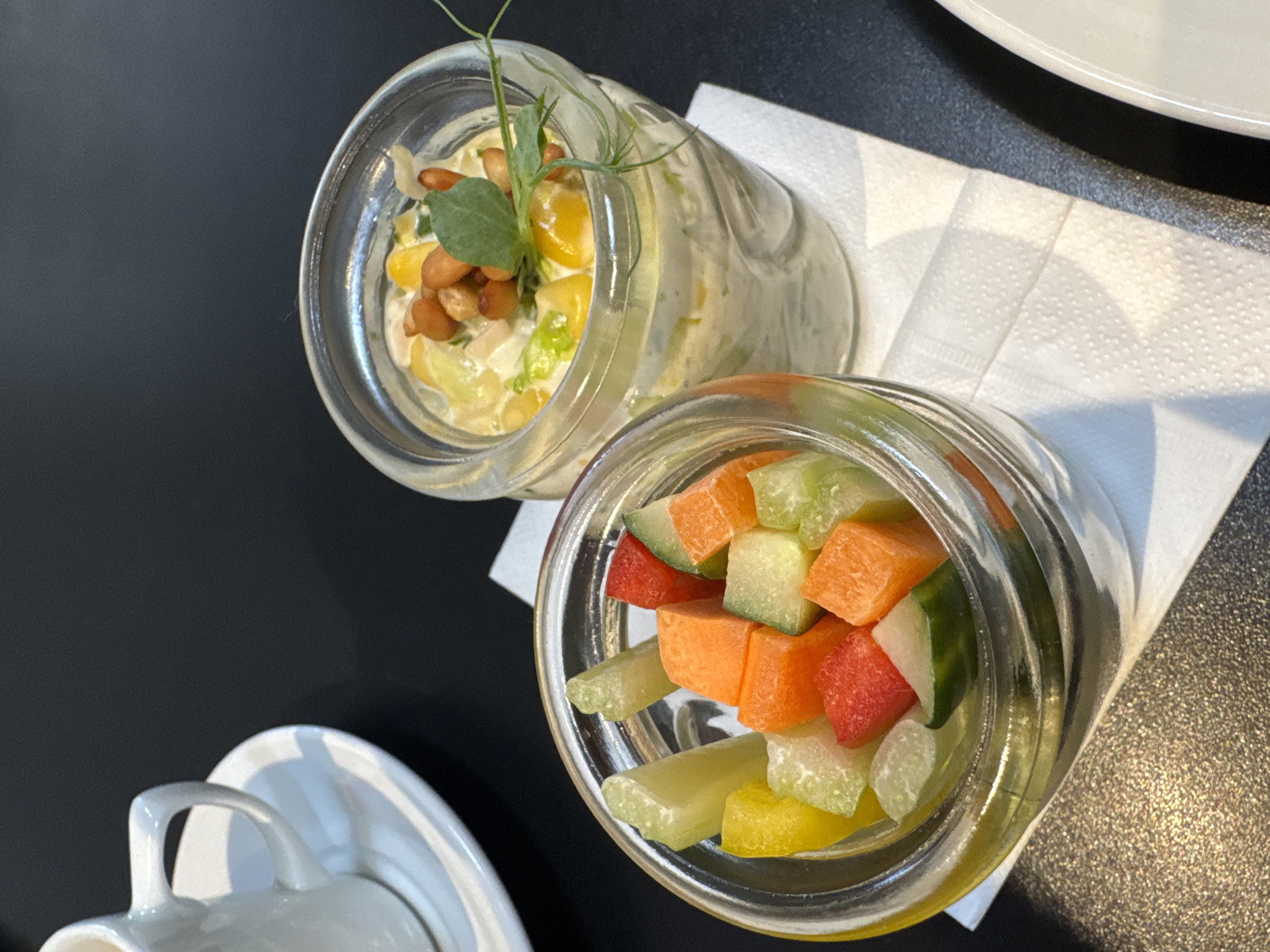 A high-angle shot reveals two glass jars containing food, resting on a white napkin on a textured, dark surface. In the lower left corner, part of a white cup and saucer are visible. The jar on the right contains what appears to be diced vegetables, including orange (carrot), red (radish or tomato), various shades of green (cucumber and celery), and yellow. The jar on the left contains a creamy salad with corn kernels and possibly peanuts, garnished with greens. The setting appears to be indoors, likely a restaurant.