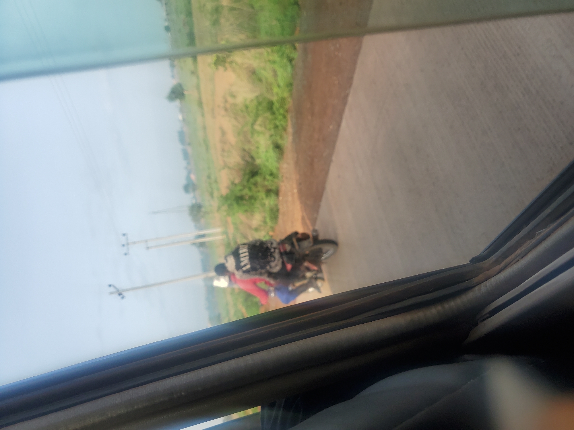 Two individuals are riding a motorcycle on a paved road. The person in the back is wearing a backpack with the word "AMIR" printed on it. The scene appears to be in a rural or semi-rural area, with fields and scattered vegetation visible in the background. The sky is clear and light. The photo is taken from inside a vehicle, as evidenced by the presence of a curtain or car interior in the foreground and the slightly distorted perspective. This image was captured in Kakau, Nigeria.